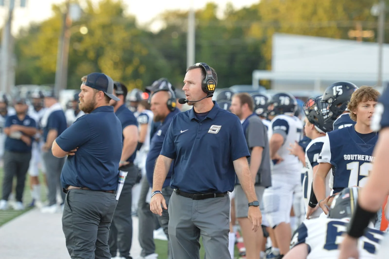 Football coaches and players standing on the sideline during a game, wearing navy blue and white uniforms, with some players wearing helmets, on a football field.