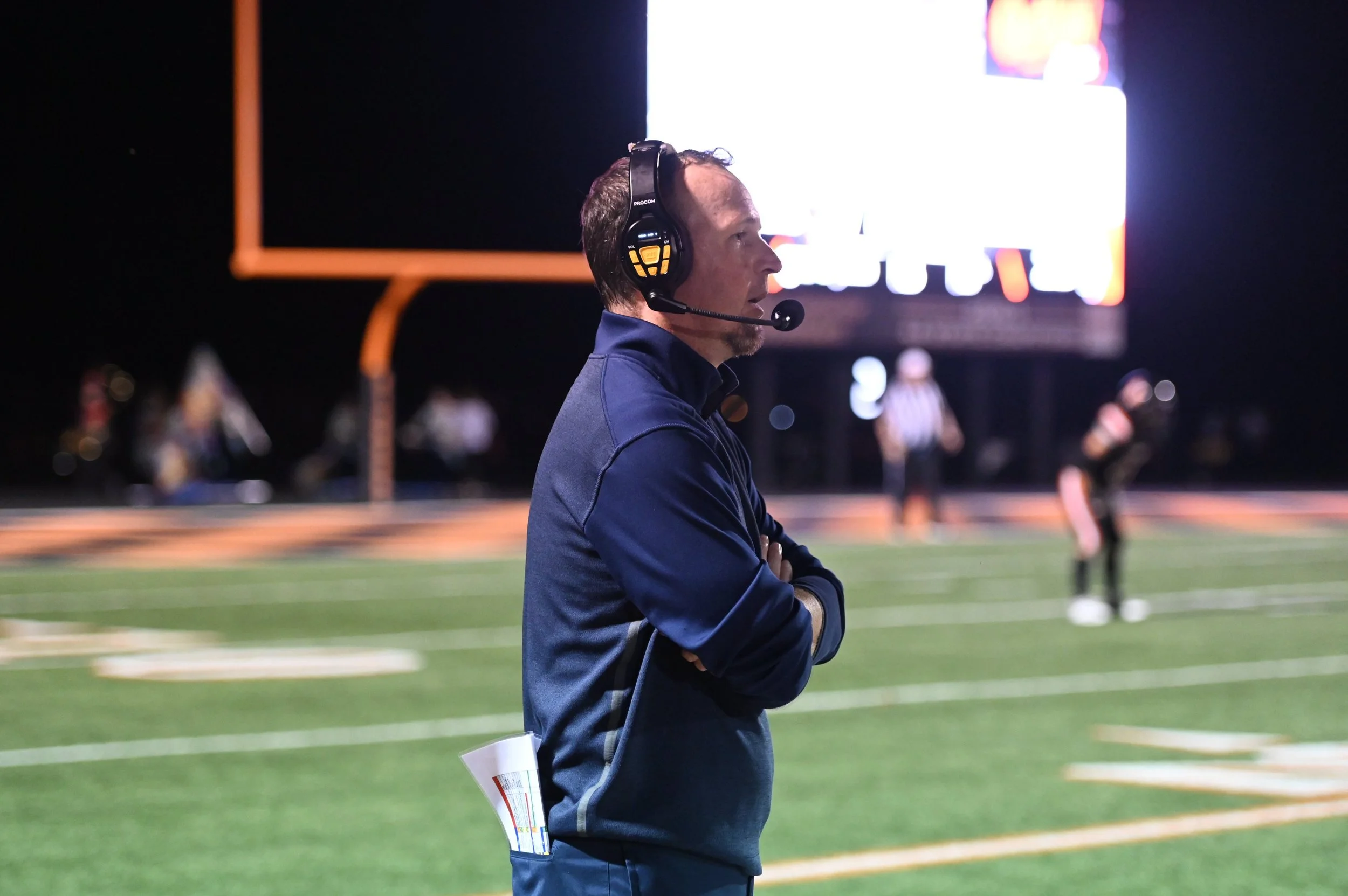 A football coach wearing a headset stands with crossed arms on the sideline of a nighttime football field, with players and a scoreboard in the background.