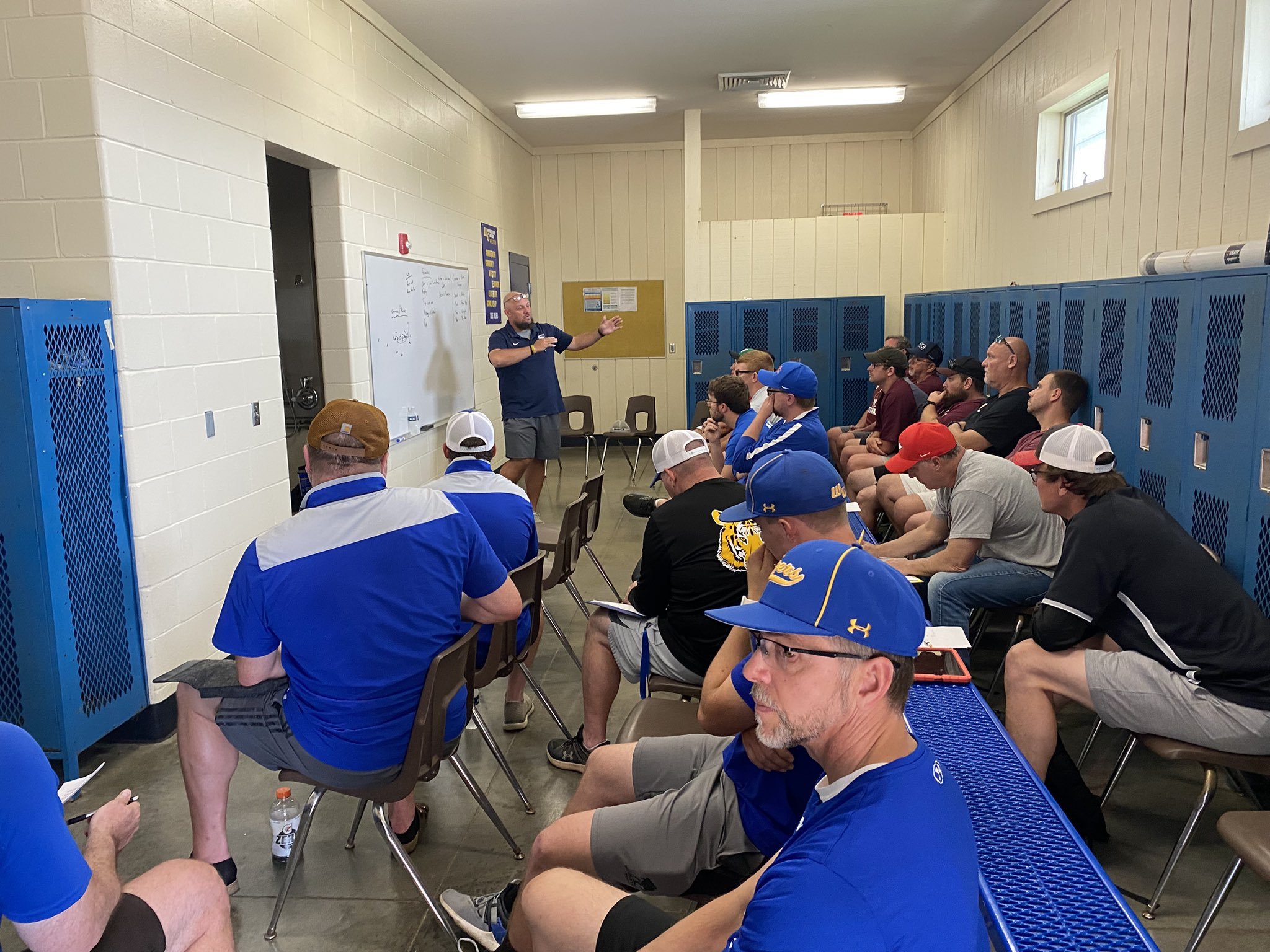 A man giving a presentation to a group of men sitting in a locker room with blue lockers, some wearing baseball caps, and a whiteboard behind the speaker.