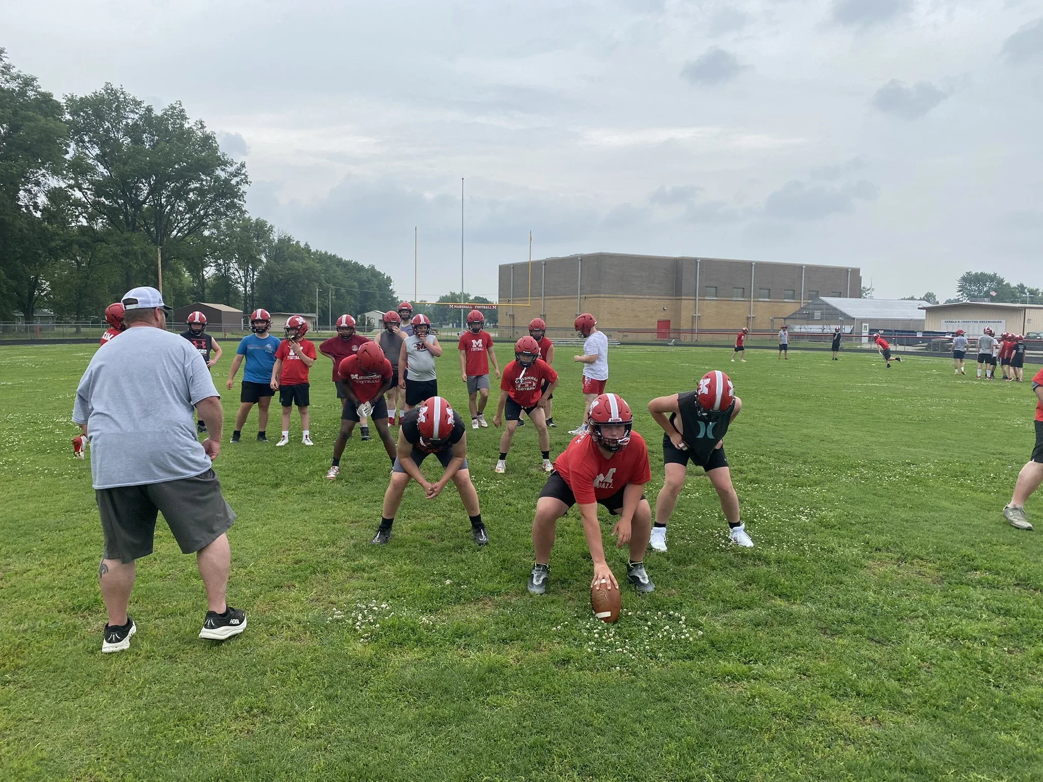Football players practicing on a green field with coaches. Players are wearing helmets and practice gear, some in red shirts. A coach in a grey shirt observes, with a large school building and trees in the background.
