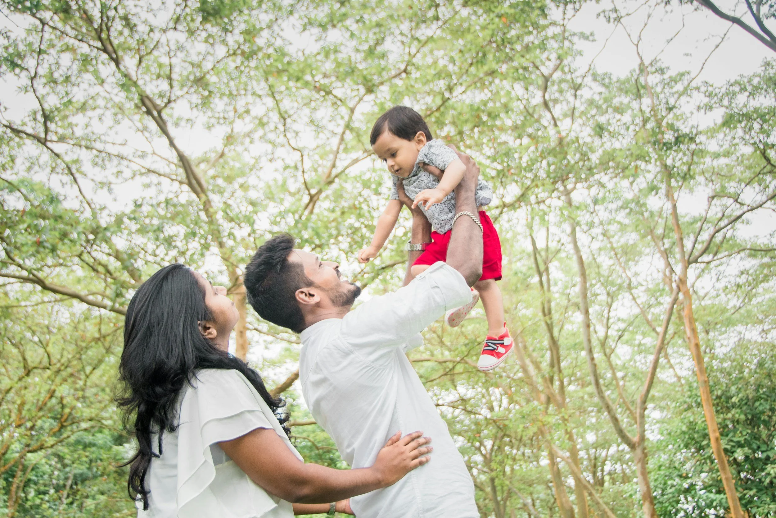 A family of three enjoying time outdoors among trees. The father lifts a young boy into the air while the mother stands beside them, both looking at the boy with smiles.