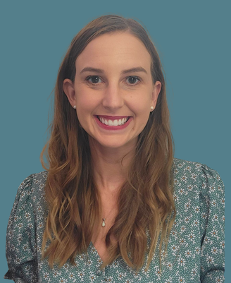 A woman with long, wavy brown hair and a big smile, wearing a teal patterned blouse and small earrings, posed against a blue background.