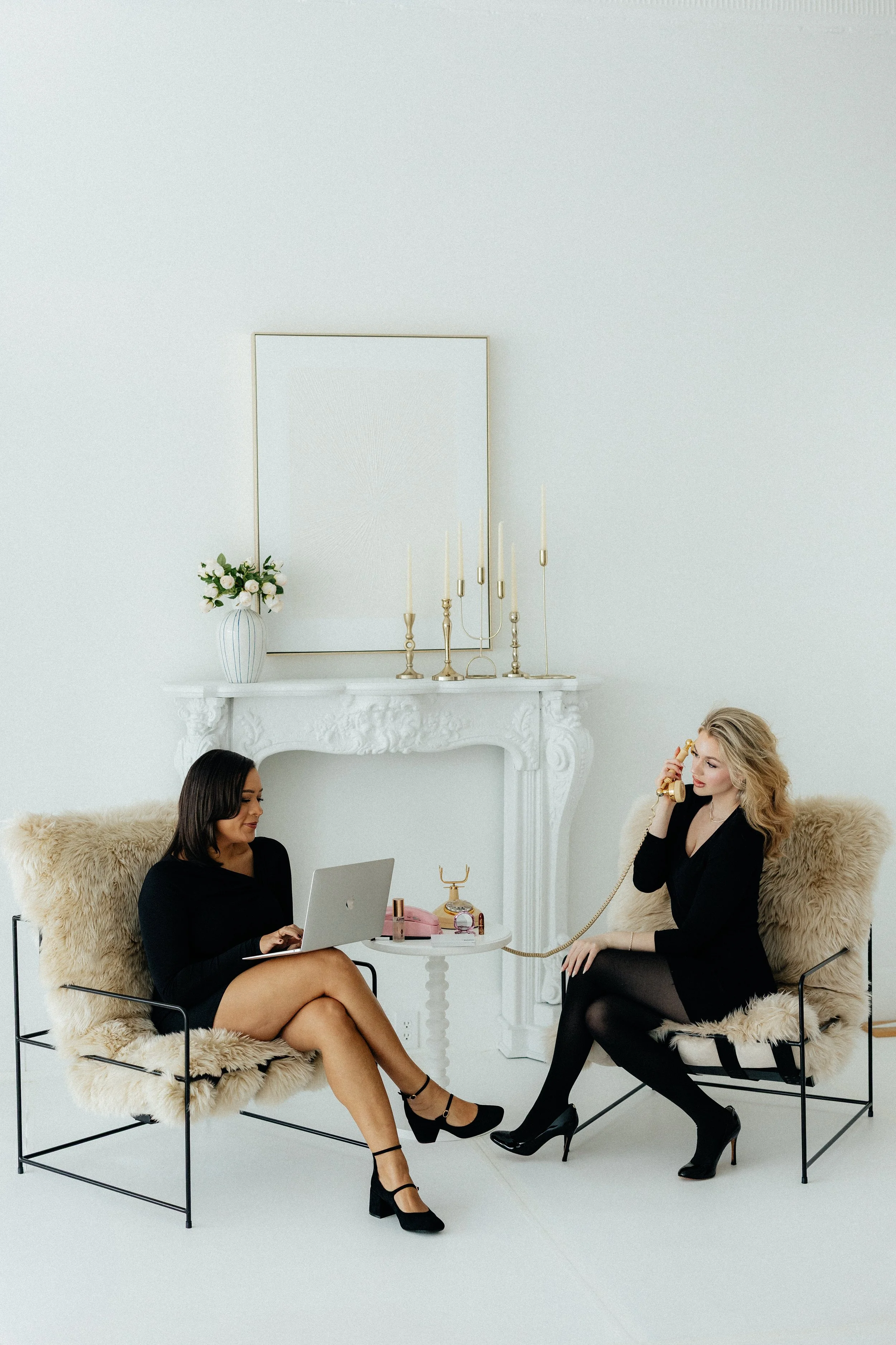 Two women sitting in a minimalist white room, one with a laptop, the other on a vintage telephone, both dressed in black, with fur-covered chairs and elegant decor.