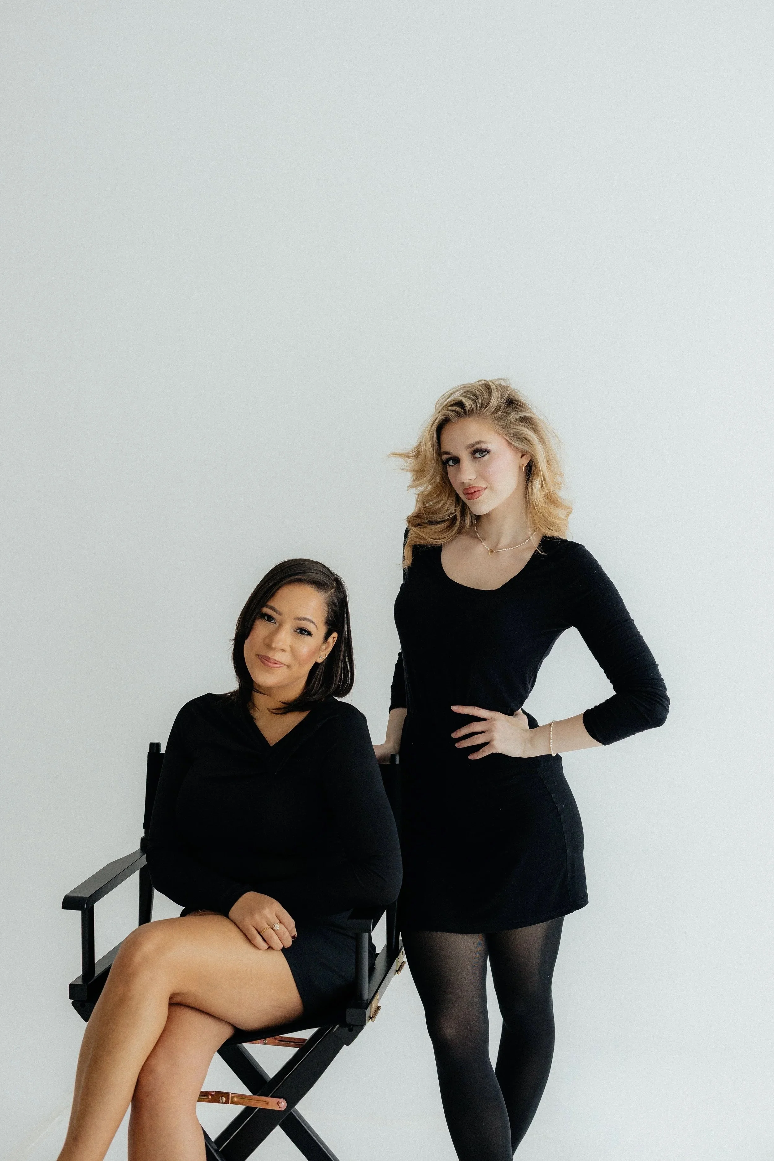Two women, one seated and one standing, posing together in front of a plain white wall. Both are dressed in black and looking at the camera.