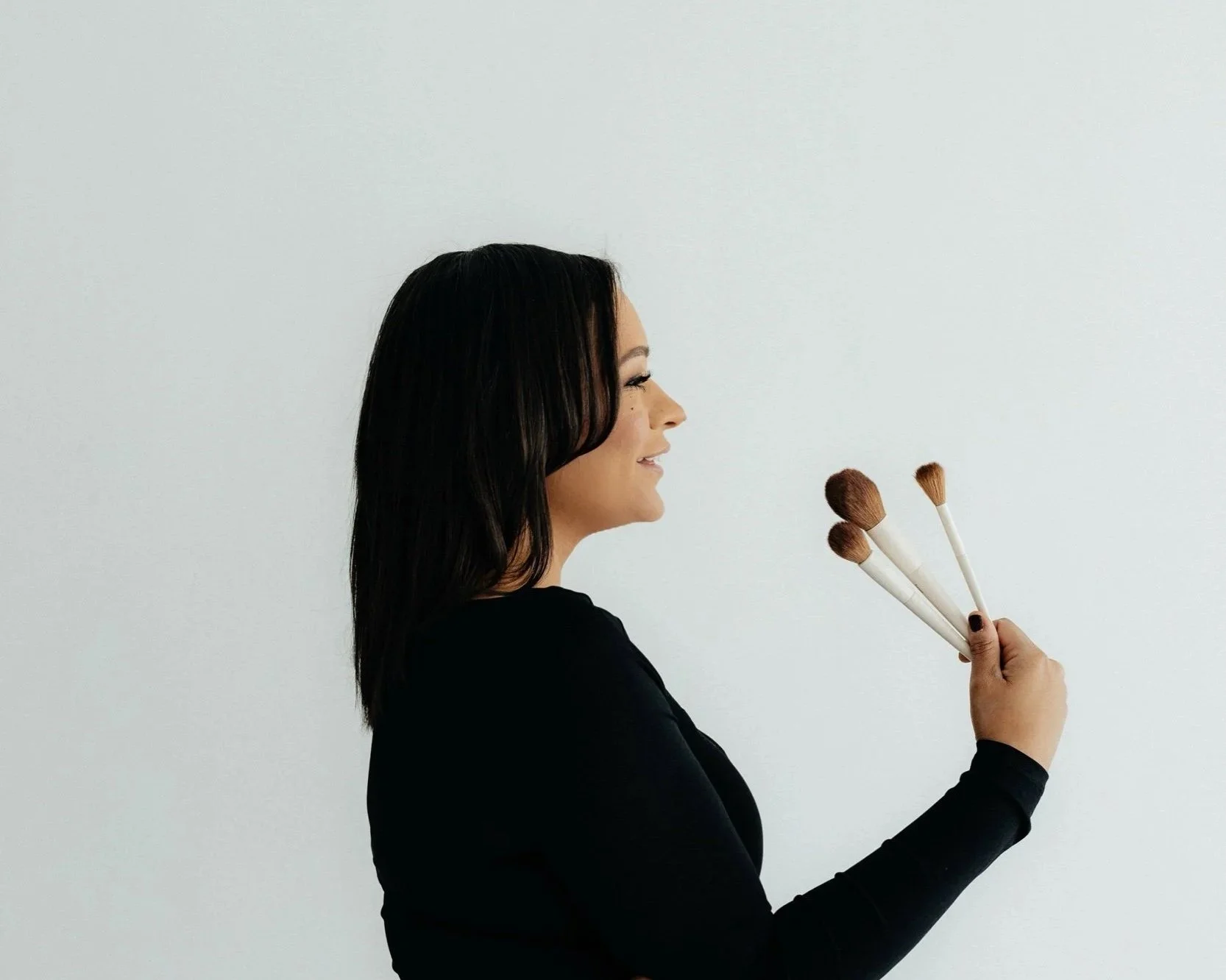 A woman with dark hair smiling and holding three makeup brushes against a plain white wall.
