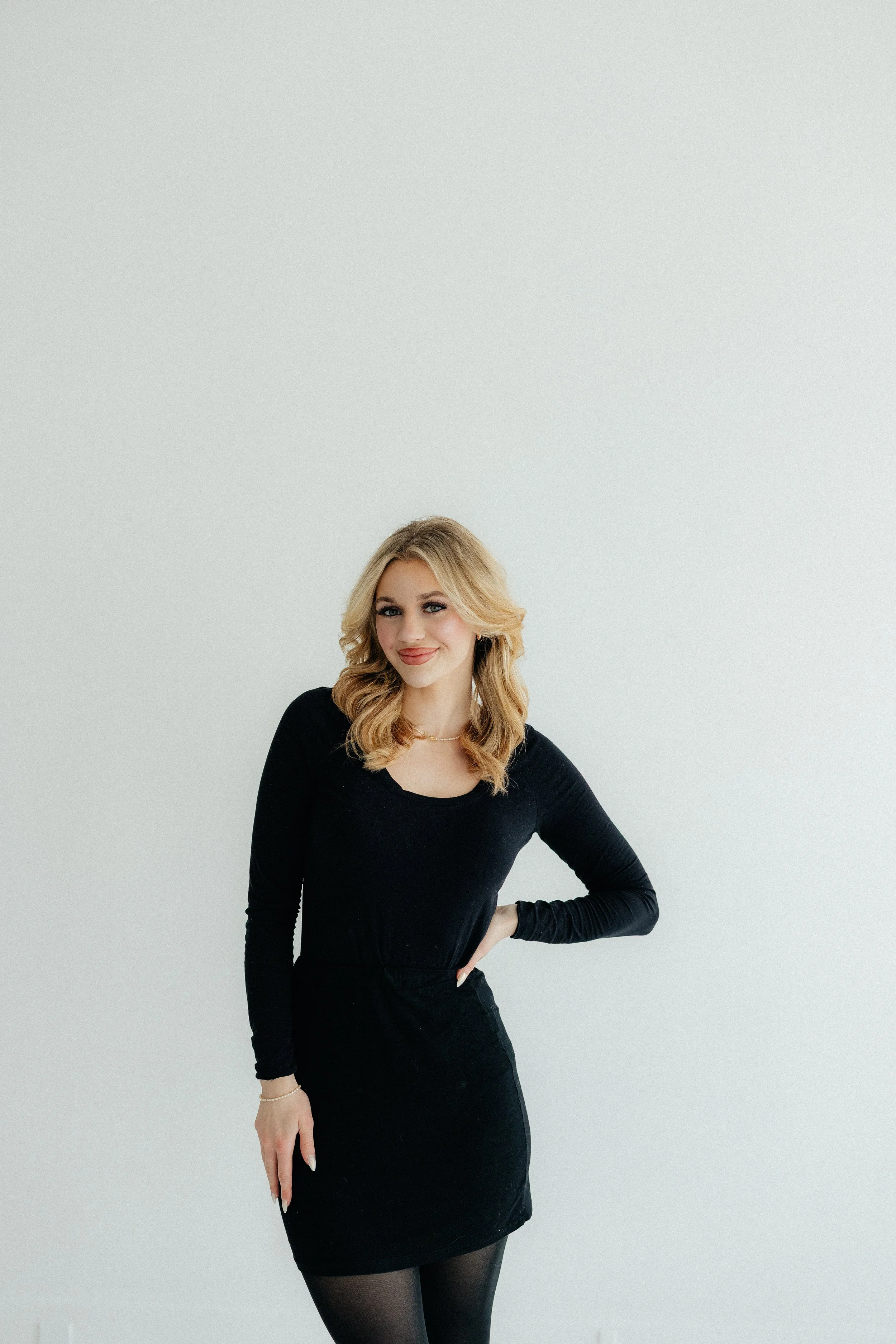 A young woman with blonde hair, wearing a black long-sleeve dress and black tights, standing against a plain light background, smiling with her hand on her hip.