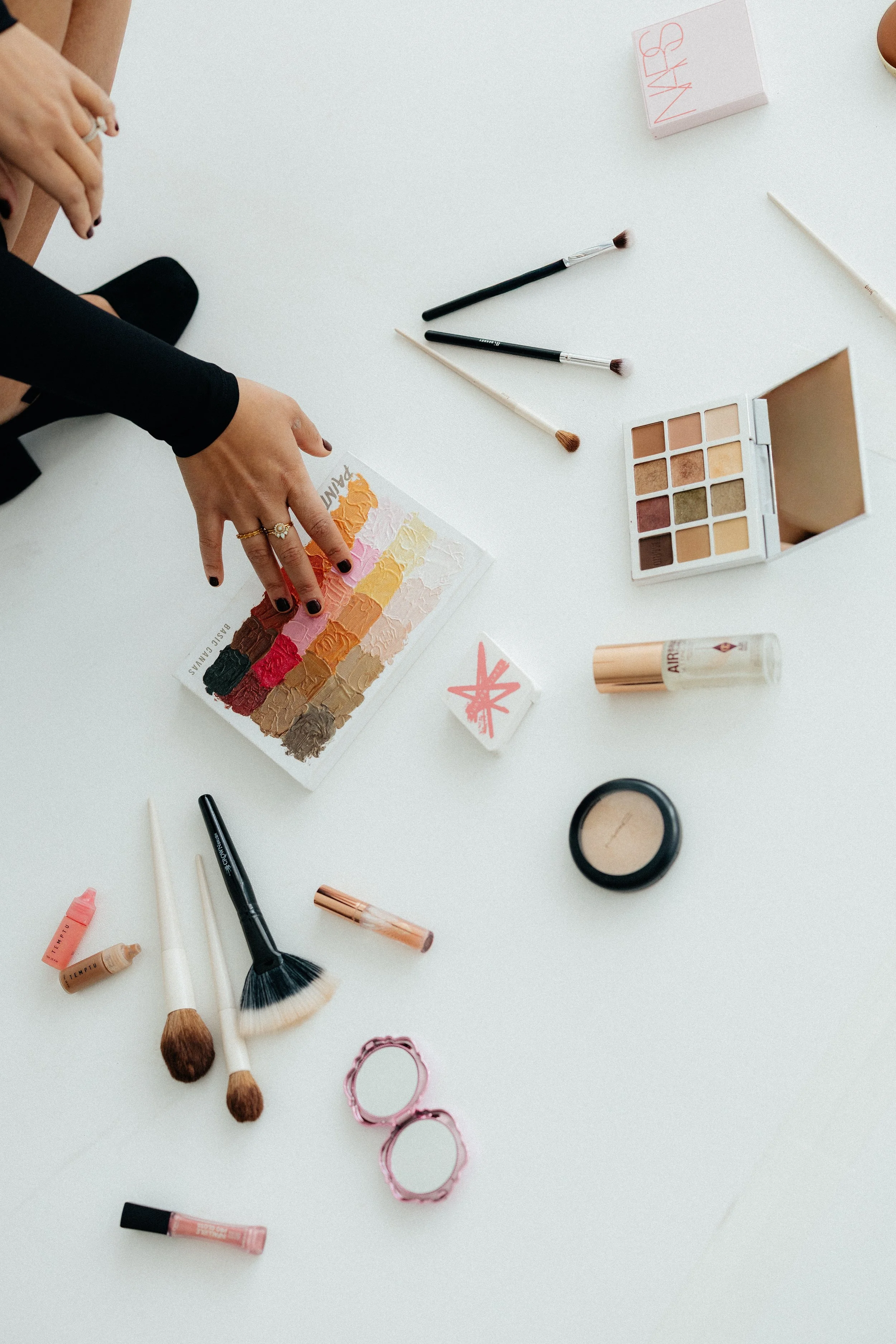 A person's hands reaching for a colorful eyeshadow palette on a white table, surrounded by makeup brushes, lipstick, compact, and other makeup products.