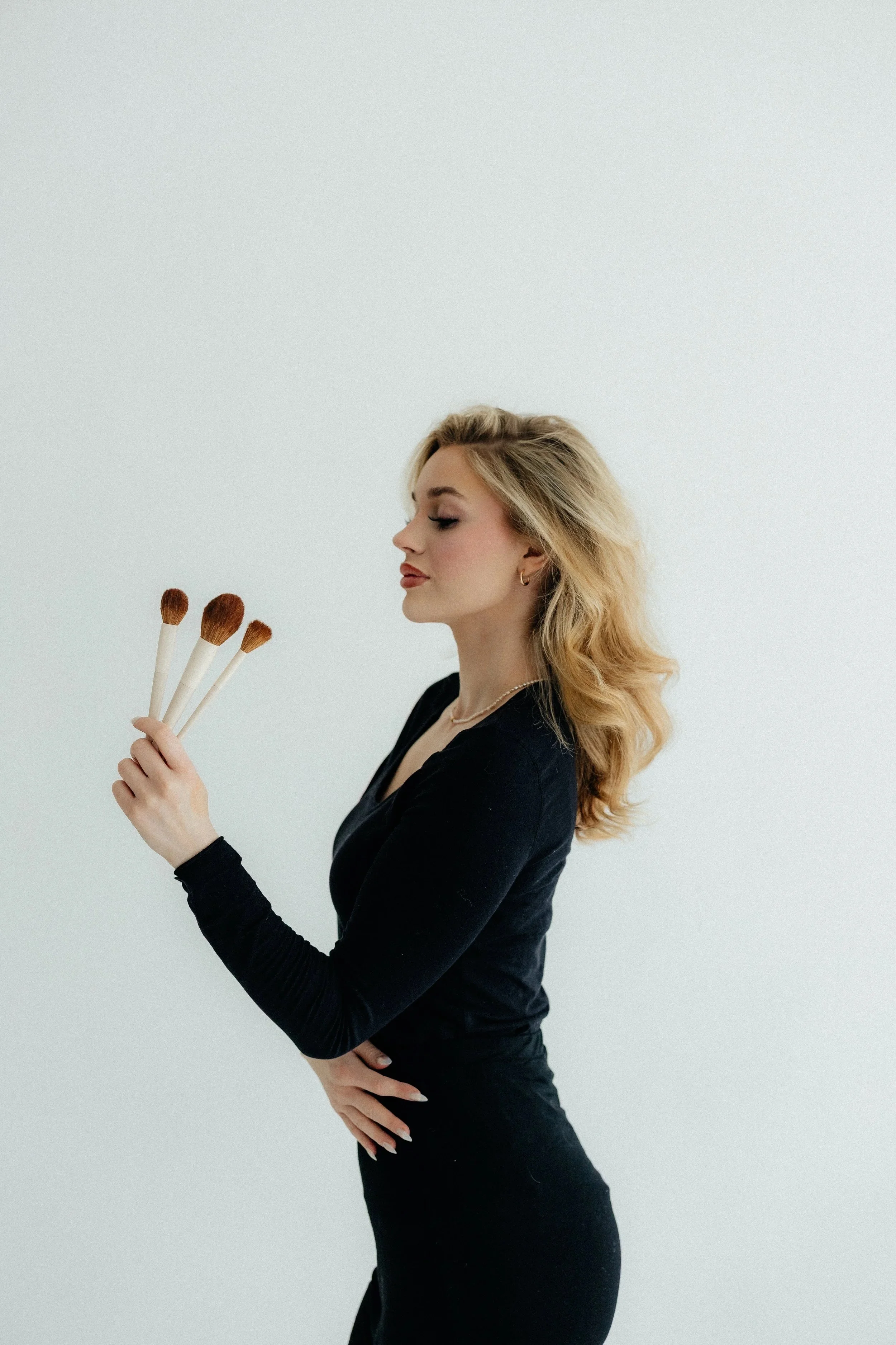 A woman with blonde wavy hair in a black long-sleeve dress holding four makeup brushes in her right hand, standing against a plain white background.