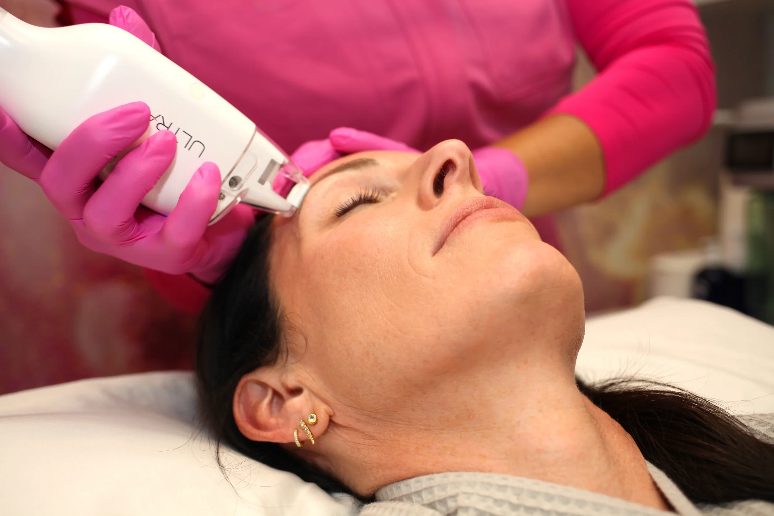 A woman with dark hair and gold earrings lying on a bed or table with her eyes closed while a person in pink gloves applies a facial treatment with a device.