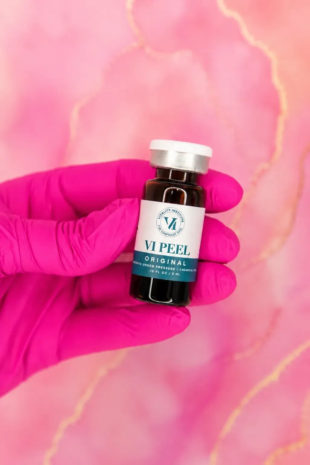 A hand wearing a pink glove holding a small bottle labeled 'VI Peel Original' in front of a pink marbled background.