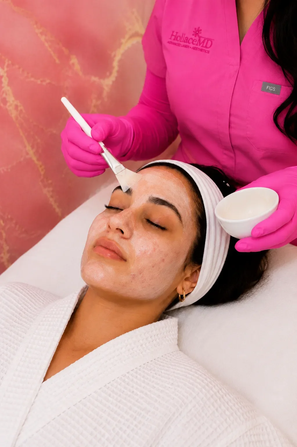A woman lying on a treatment bed with her eyes closed, receiving a facial treatment from a skincare professional in a pink uniform and pink gloves. The professional is applying a facial product with a brush.