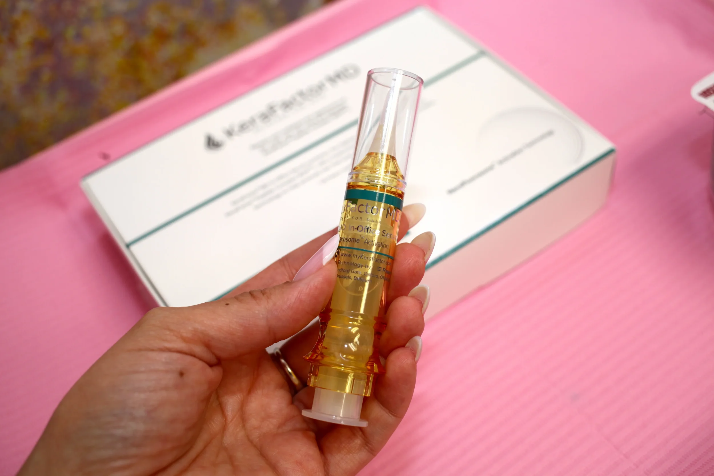 A person holding a yellow-colored medical syringe with a clear plastic needle cap, with a box in the background on a pink surface.
