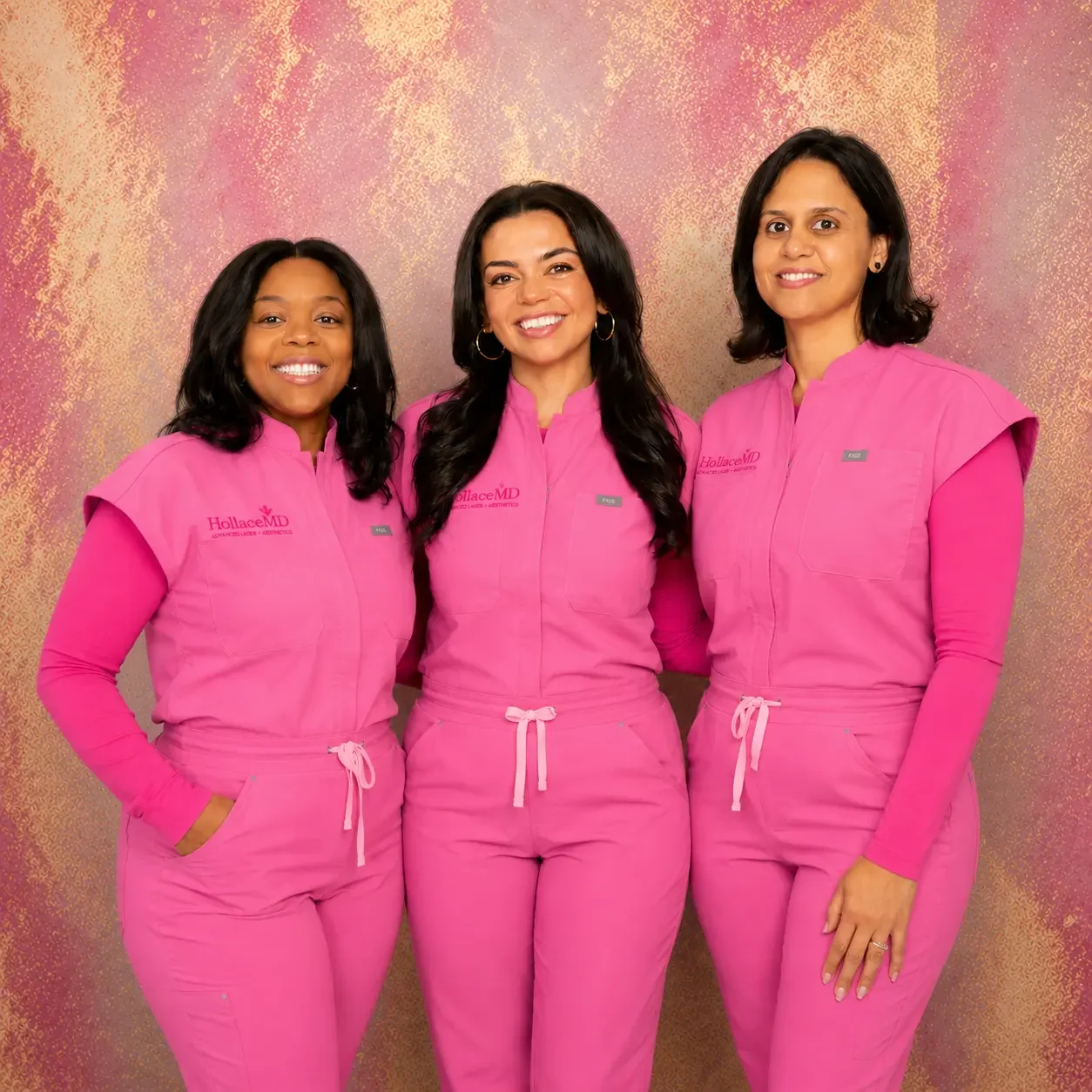 Three women standing together, smiling, wearing matching pink medical scrubs with 'Hollace MD' on the left chest, against a pink and gold textured background.