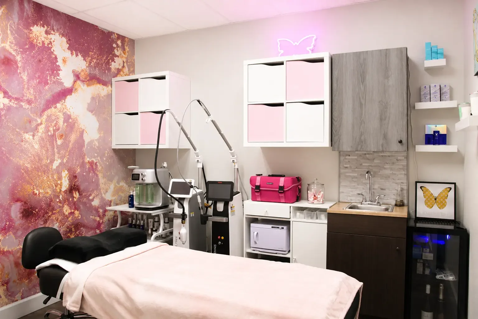 Interior of a medical or spa treatment room with a treatment bed covered in pink, surrounded by medical equipment, cabinets, and decor in soft pastel and pink tones. A pink neon butterfly light is on the wall.