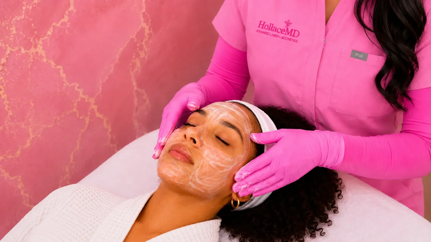 A woman receives a facial treatment at a spa, lying with eyes closed while a skincare specialist applies a facial mask. The specialist wears pink gloves and a pink uniform with the logo 'HollaceMD' on it.