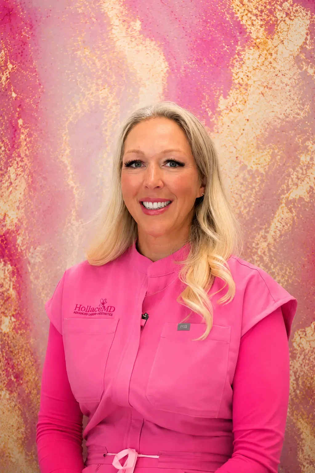 A woman with long blonde hair smiling, wearing a bright pink medical uniform with the logo 'HollaceMD Advanced Laser Aesthetics,' standing in front of a pink and gold abstract background.