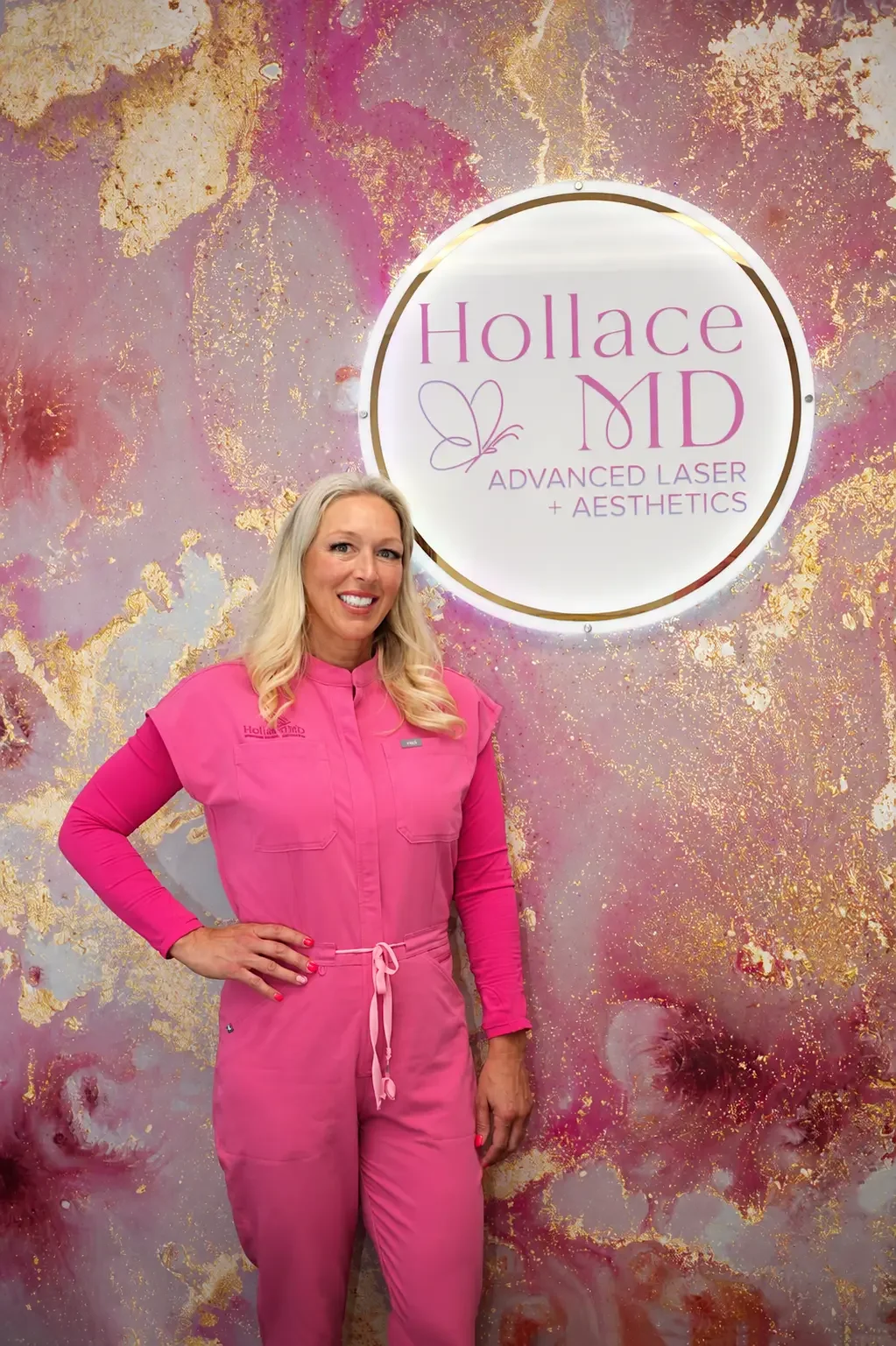 Woman with long blonde hair smiling, wearing a bright pink medical uniform, standing in front of a vibrant pink, purple, and gold abstract background and a circular sign that reads 'Hollace MD Advanced Laser + Aesthetics' with a butterfly logo.