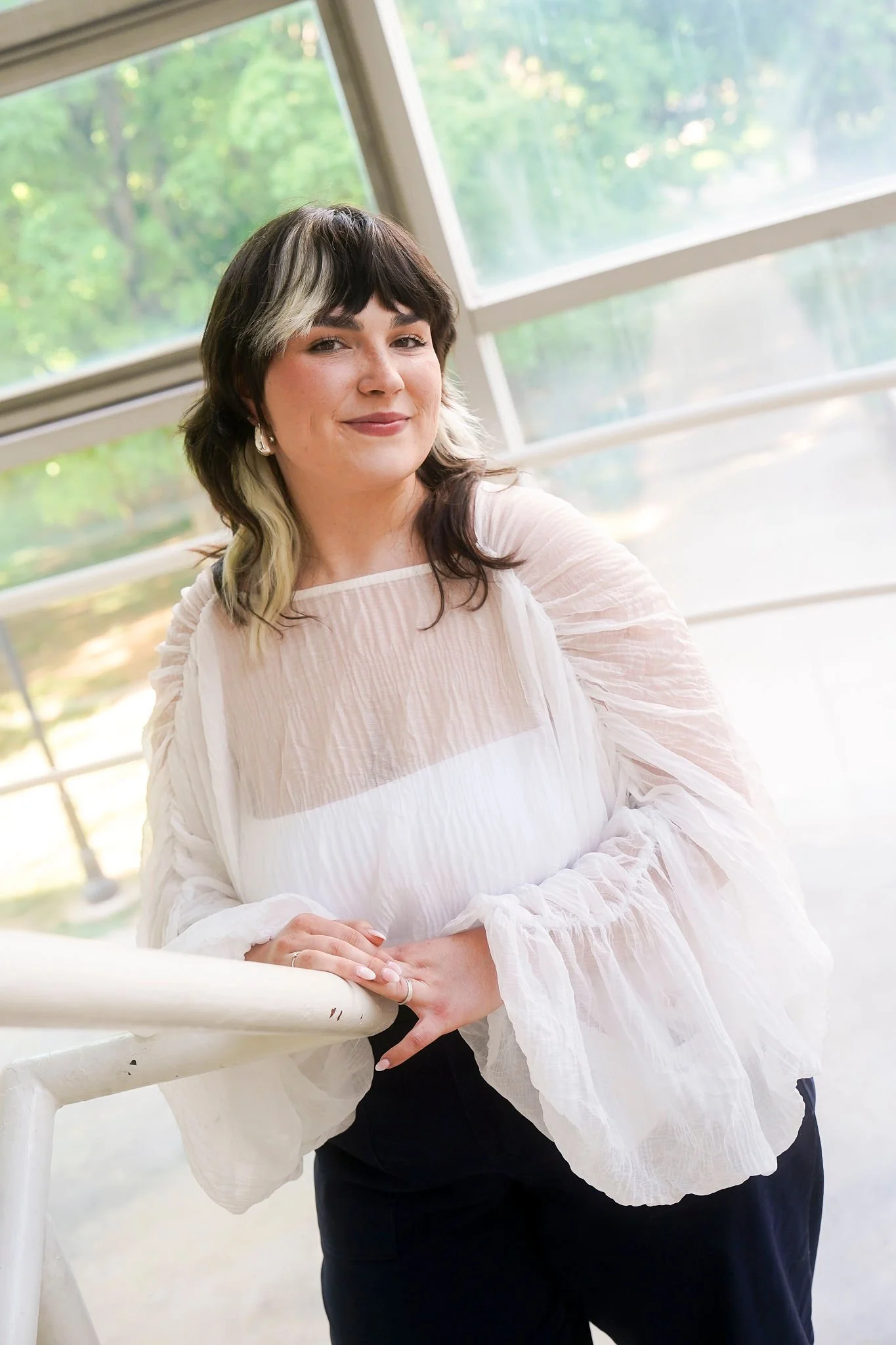 A woman with dark hair streaked with blonde, wearing a white blouse with ruffled sleeves, leaning on a white railing with her hands clasped, in front of large windows with a green outdoor scene visible.