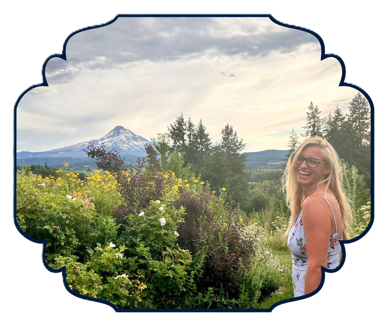 A woman with blonde hair and glasses smiling in a floral dress in a garden with colorful flowers and greenery, with a mountain and cloudy sky in the background.