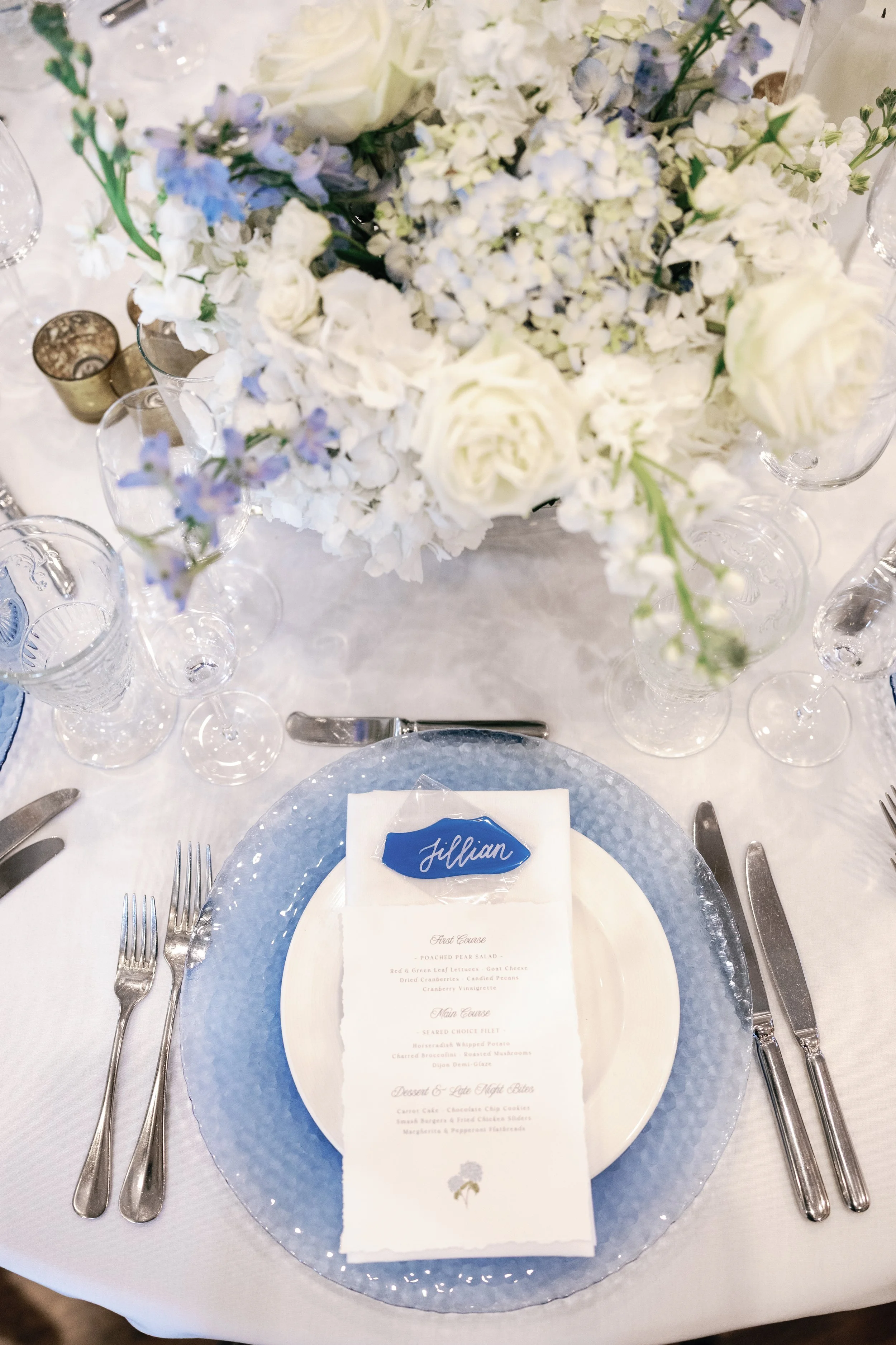 Elegant table setting with a hand-lettered sea glass place card in white ink, floral centerpiece of white and light purple flowers, silverware, glassware, and a menu on a blue plate with a name tag labeled 'Jillian' at a formal event.