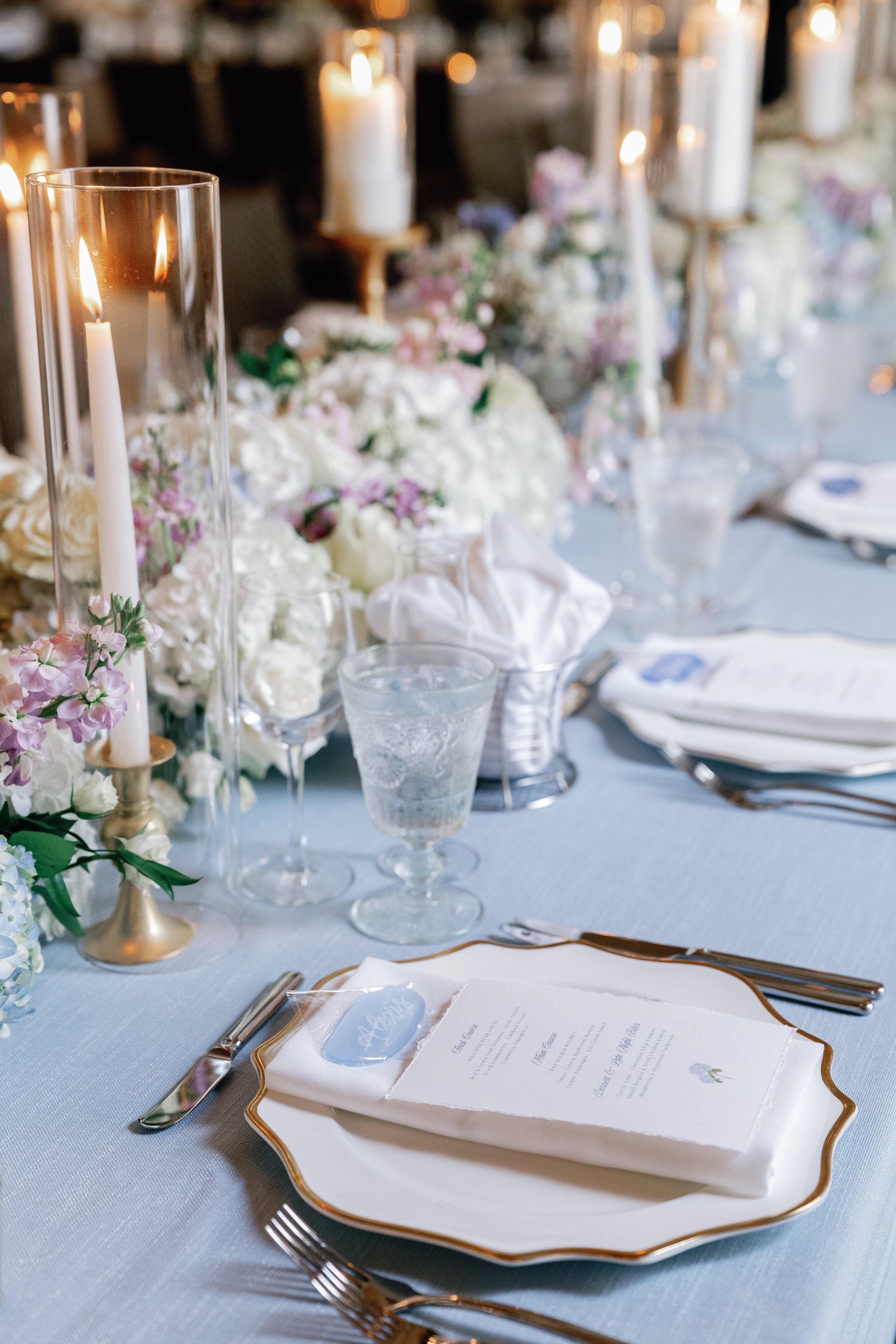 Elegant wedding reception table setting with white floral centerpiece, tall candles in glass holders, water glasses, plates with gold trim, and silverware, with a hand-torn dinner menu and a blue sea glass place card