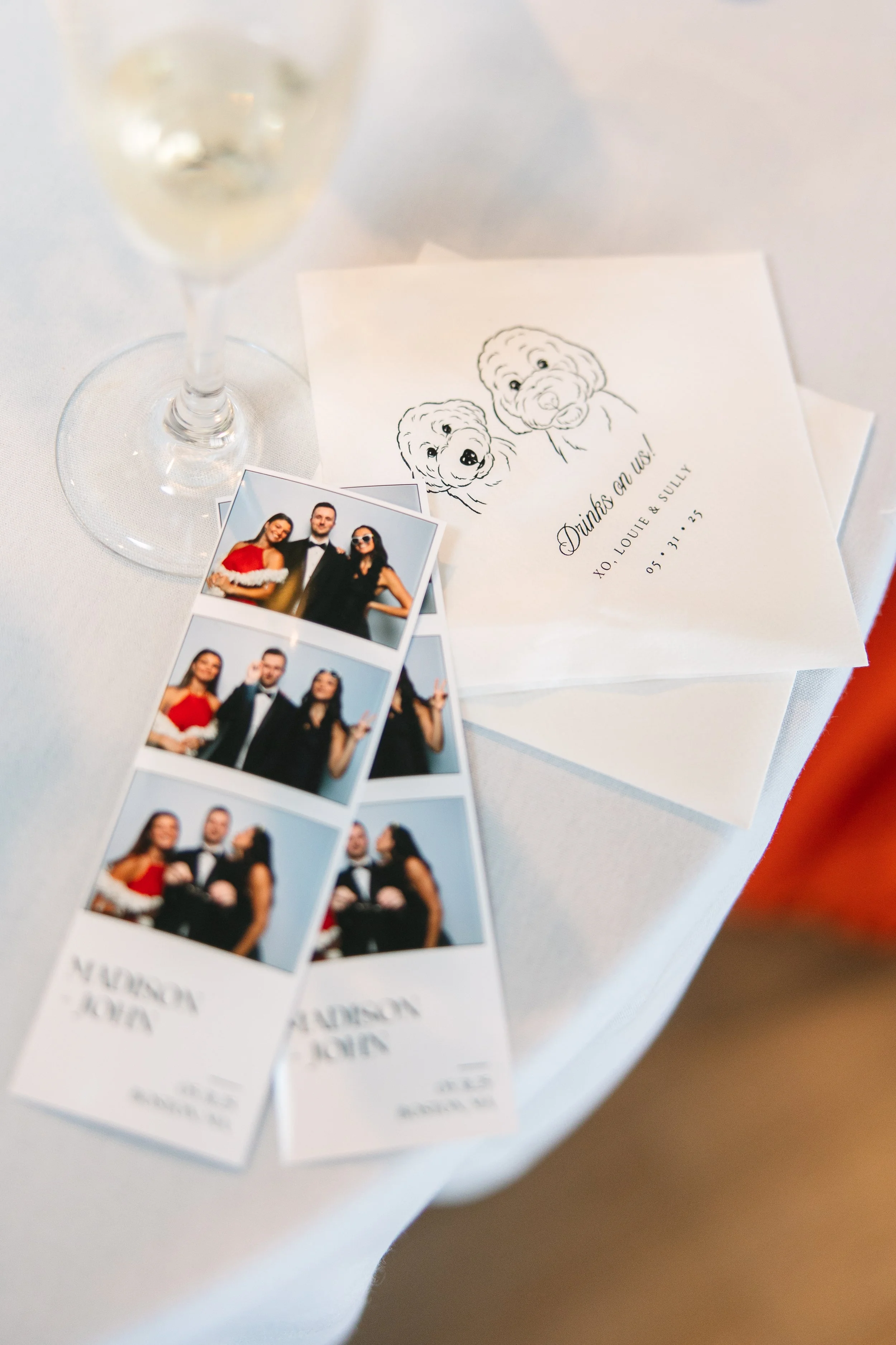A table with photo booth pictures, a glass of wine, and a cocktail napkin with dog portrait and the message 'Drinks on us! XO Louie & Sully 05.31.23'.