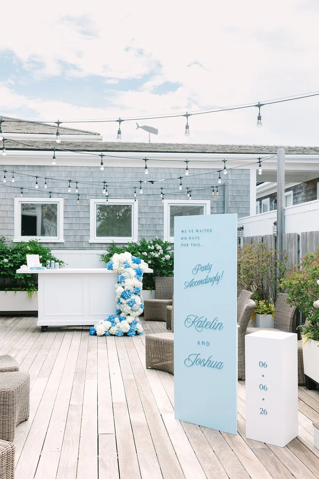 Nantucket wedding celebration setup with a large blue welcome sign, white pedestal with wedding date, floral arrangements, string lights, and a small bar laced with hydrangeas