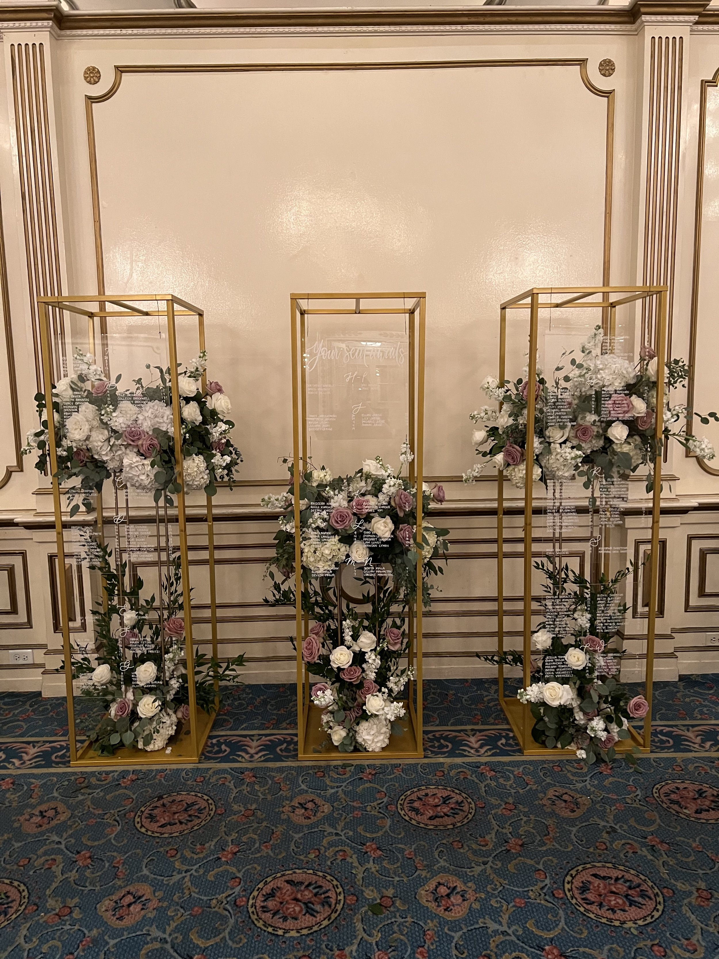 Three gold metal stands with clear acrylic seating chart and floral arrangements of white and purple roses, hydrangeas, and greenery, arranged in a decorative display against a cream wall with gold accents at the Fairmont Copley Plaza