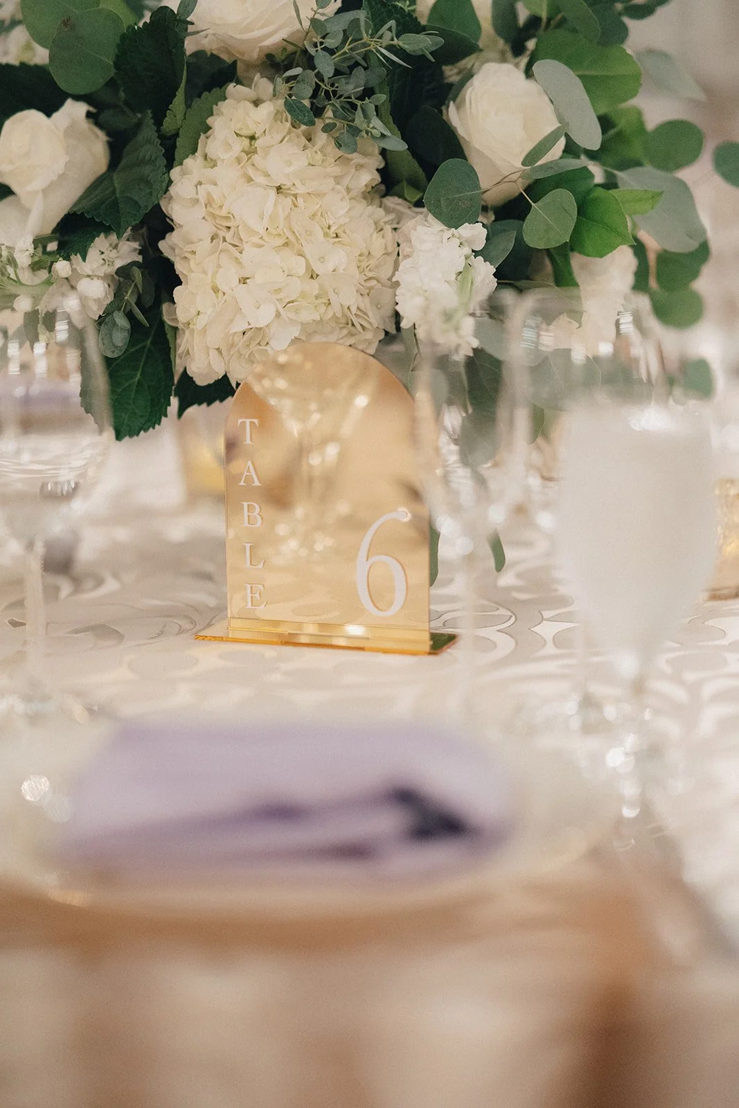Wedding tablescape with a gold mirror table number card displaying 'Table 6,' surrounded by glassware and a floral centerpiece of white hydrangeas and greenery.