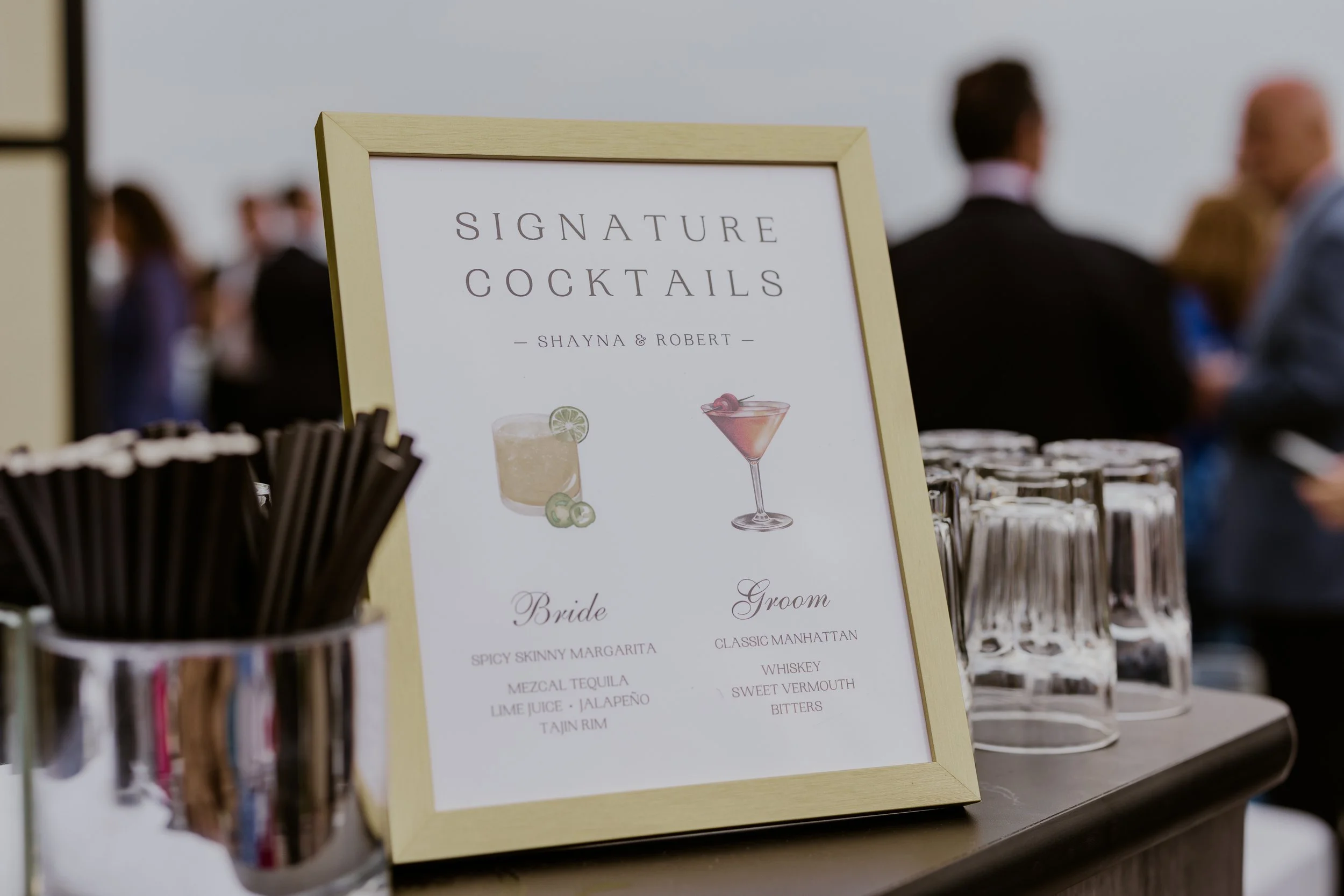 A sign on a table advertising signature cocktails at an event, with illustrations of a cocktail glass and a drink with a lime garnish, and lists of drinks for the bride and groom.