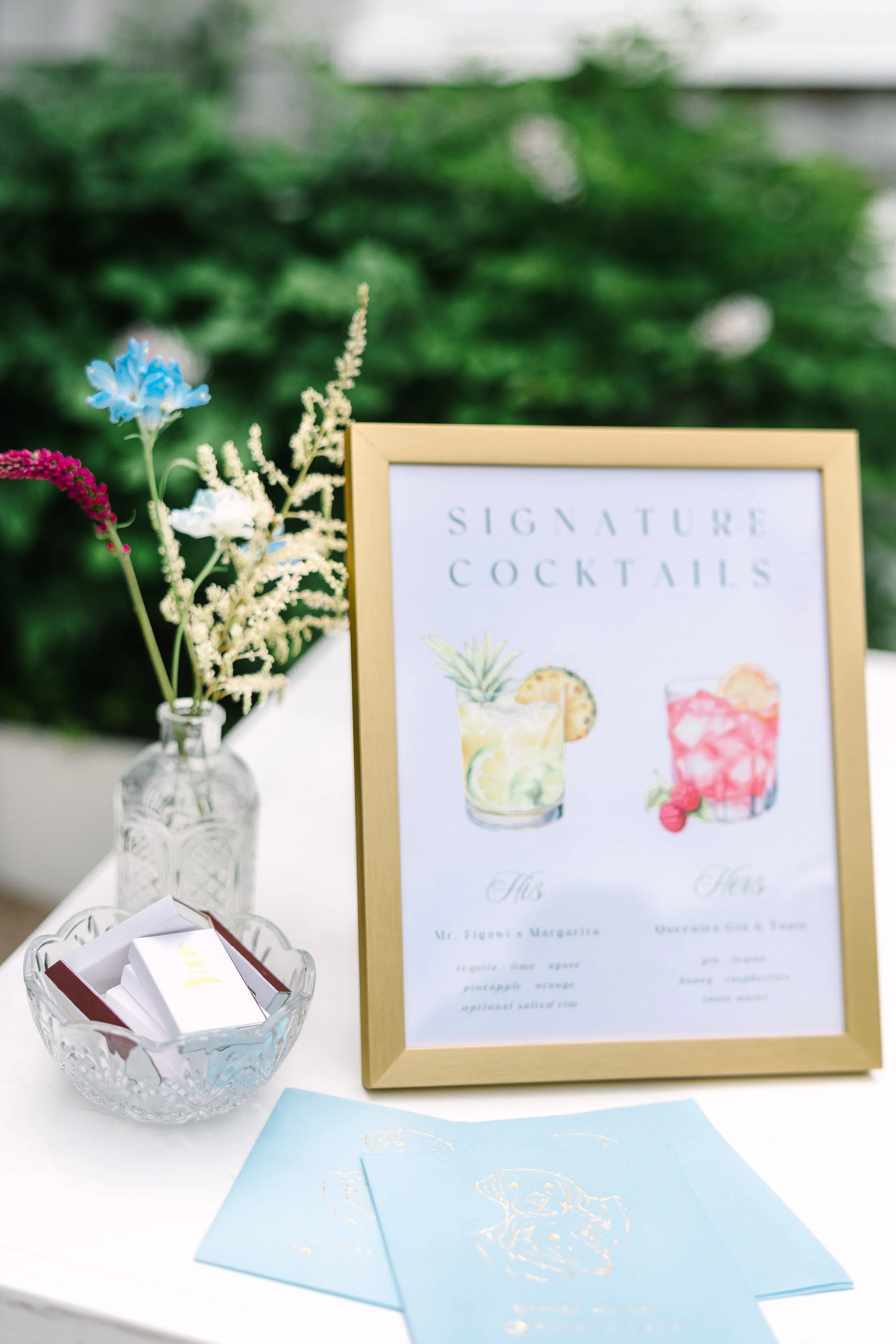 A wedding cocktail sign for signature drinks with watercolor illustrations of drinks and tropical fruits, paired with dog pet portrait napkins and lighthouse matchboxes