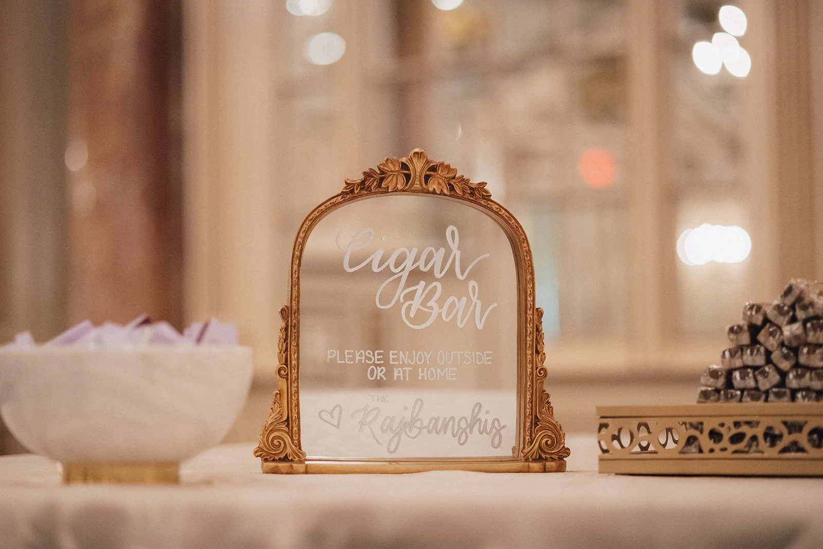Ornate wedding cigar b sign with "Cigar Bar, Please enjoy outside or at home". The sign is placed on a table with a bowl of purple match boxes on the left and a tray of cigars on the right.