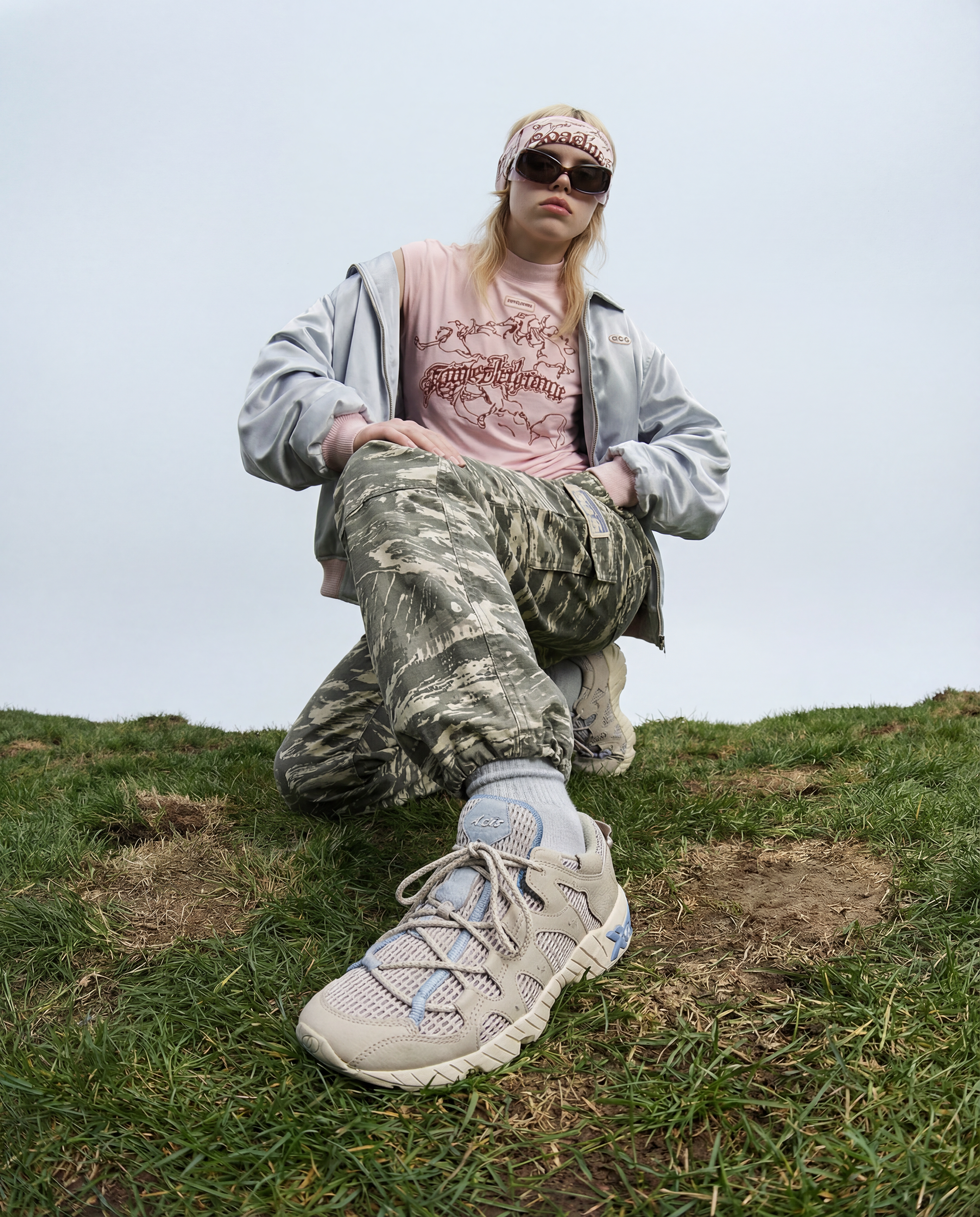 A person wearing sunglasses, a pink headband, a pink graphic t-shirt, a silver jacket, camouflage pants, and light-colored sneakers, kneeling on a grassy hill with an overcast sky in the background.