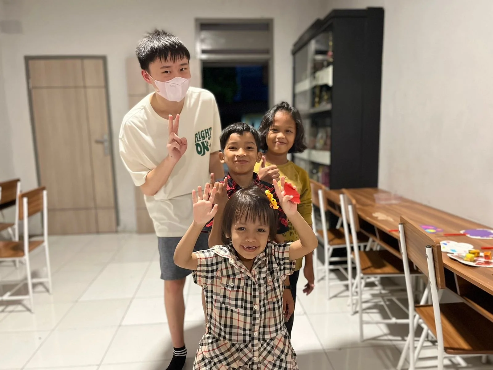 A group of four children standing indoors, smiling and waving at the camera, with a table and chairs on the side.