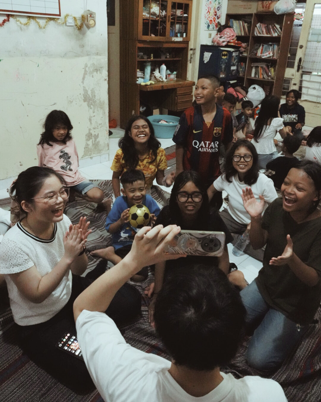 Group of children and adults smiling and laughing during a gathering indoors, some sitting on the floor and one person taking a photo with a smartphone.