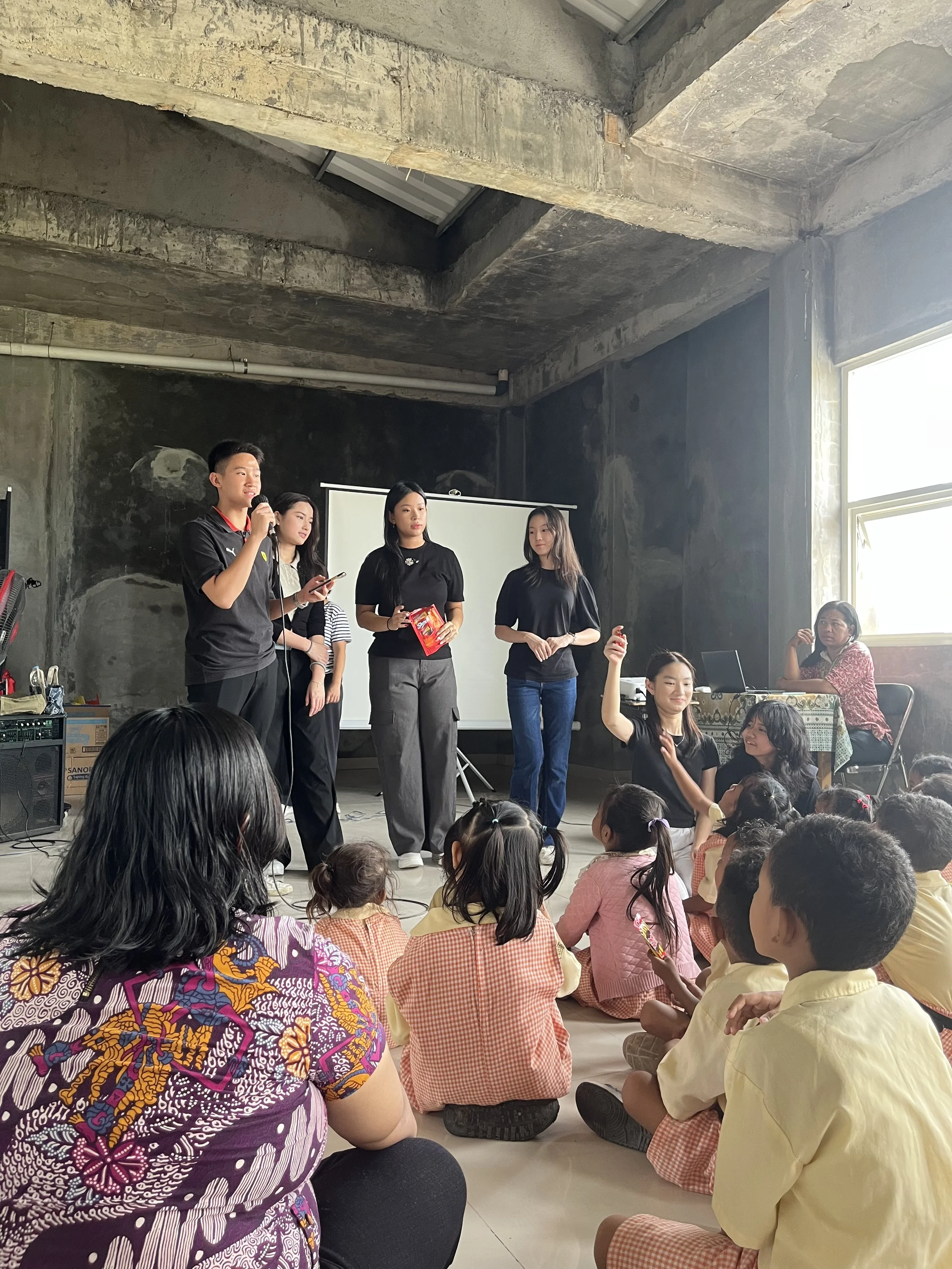 A group of young women and a young man are standing on a stage or platform in front of an audience of children. The children are sitting on the floor, facing the performers. The setting appears to be an indoor space with unfinished concrete walls and ceiling, with a large window letting in natural light. The children are wearing school uniforms, and the women look to be hosting or presenting.