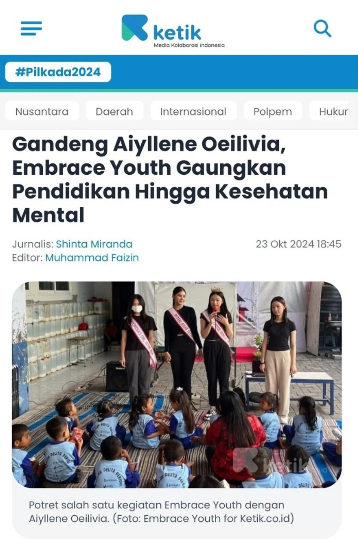 Women and children participating in Embrace Youth activity at an outdoor event in Indonesia. Women are standing and speaking to children seated on the ground, with some women wearing sashes that read 'Embrace Youth.' Children are dressed in blue uniforms and are attentively listening.