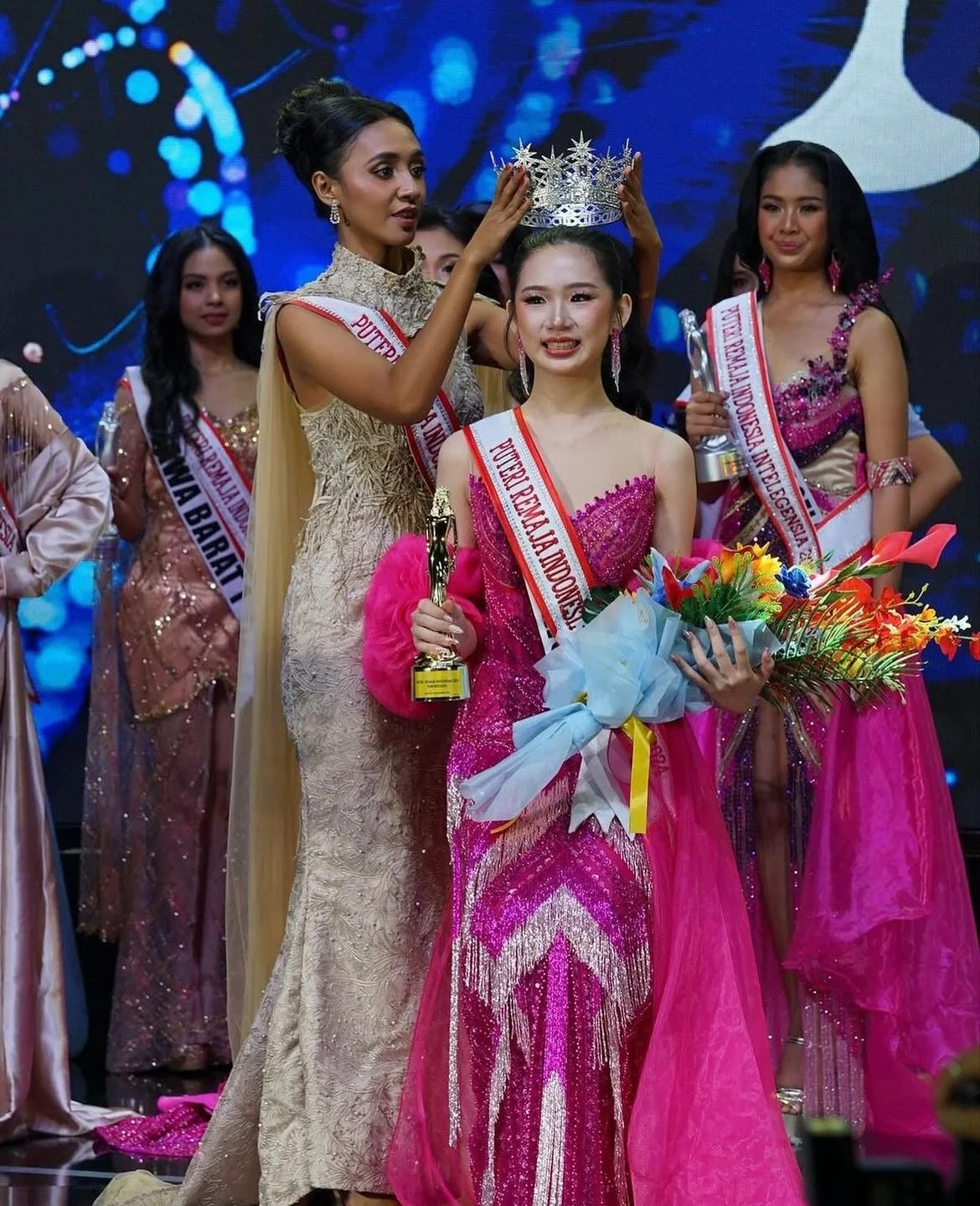 Beauty pageant winner wearing a pink dress and a sash, being crowned on stage with other contestants in evening gowns.