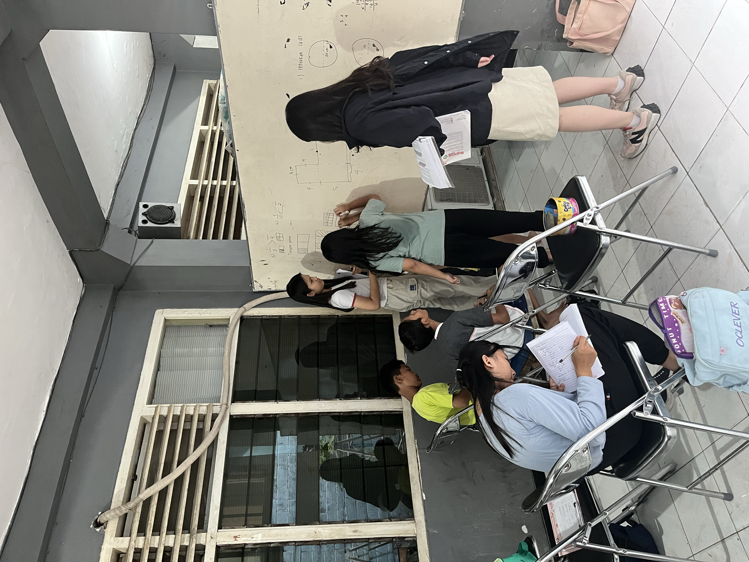 Group of students in a classroom engaging with whiteboard, some sitting at desks, others standing and writing.