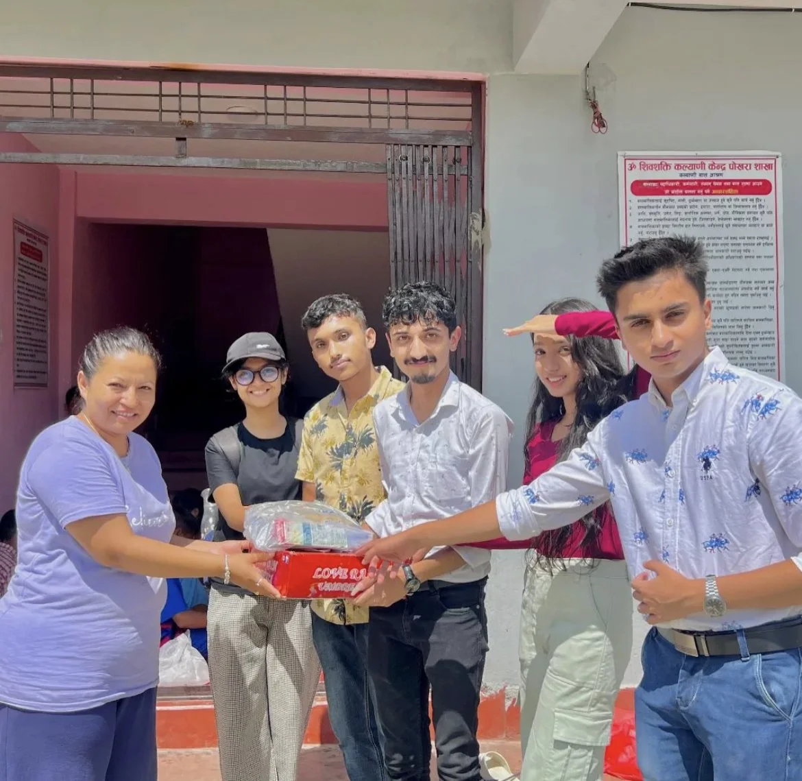 Group of six people handing over a package outside a building with a pink and gray exterior and a signboard on the wall.