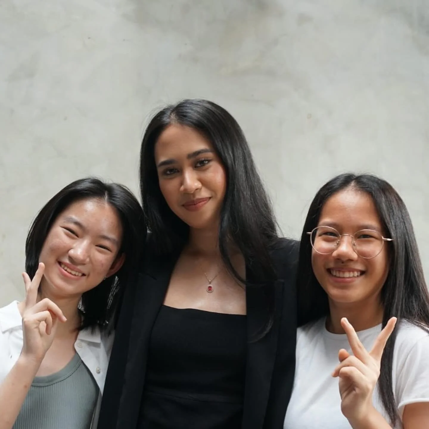 Three young women smiling and making peace signs while standing together indoors against a plain background.
