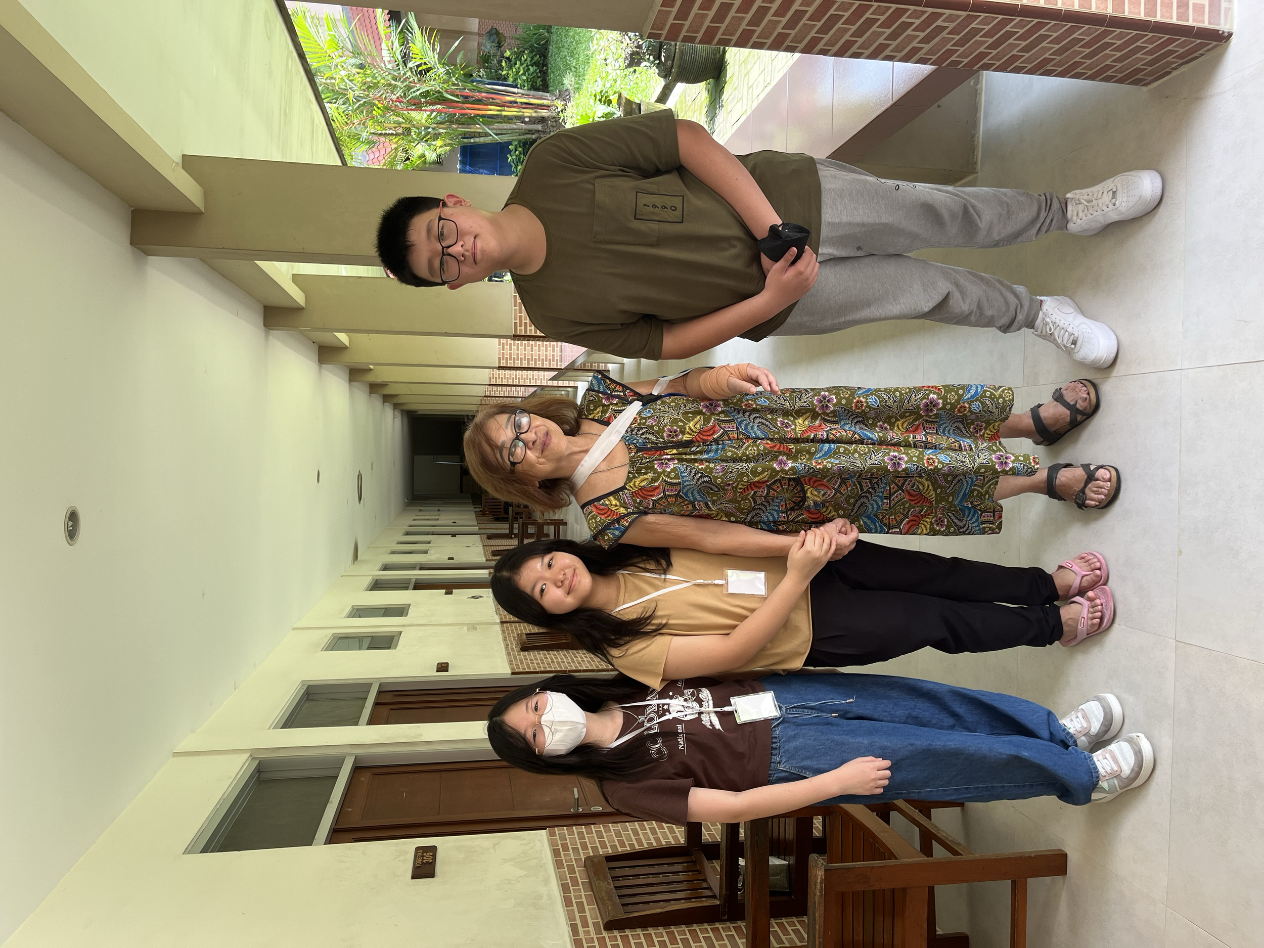 Four people standing in a hallway outdoors. Two women and one man are wearing glasses, and one woman has a face mask. They are smiling and holding hands.
