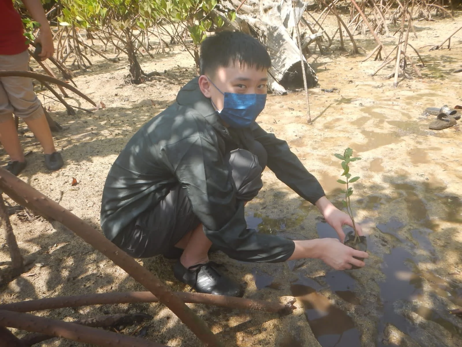 A young man wearing a blue face mask, black jacket, and black shoes, squatting down on sandy ground near water, holding a small plant in a plastic bag.