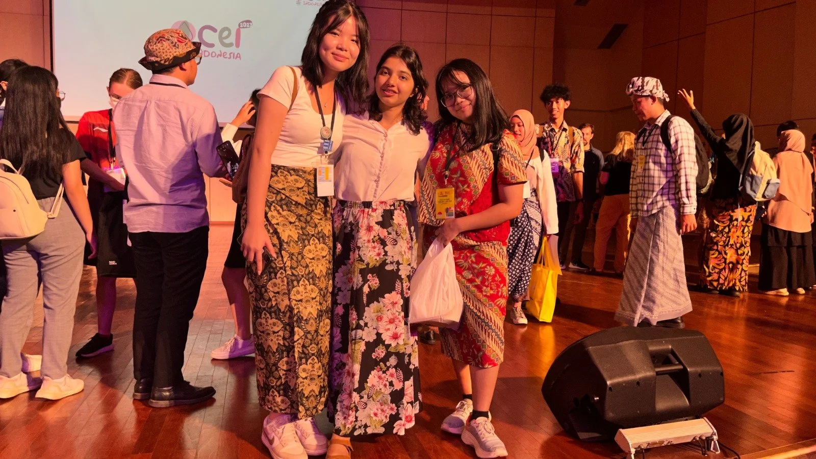 Group of young women standing together, posing for a photo at an event, with a background featuring a projector screen and other attendees.