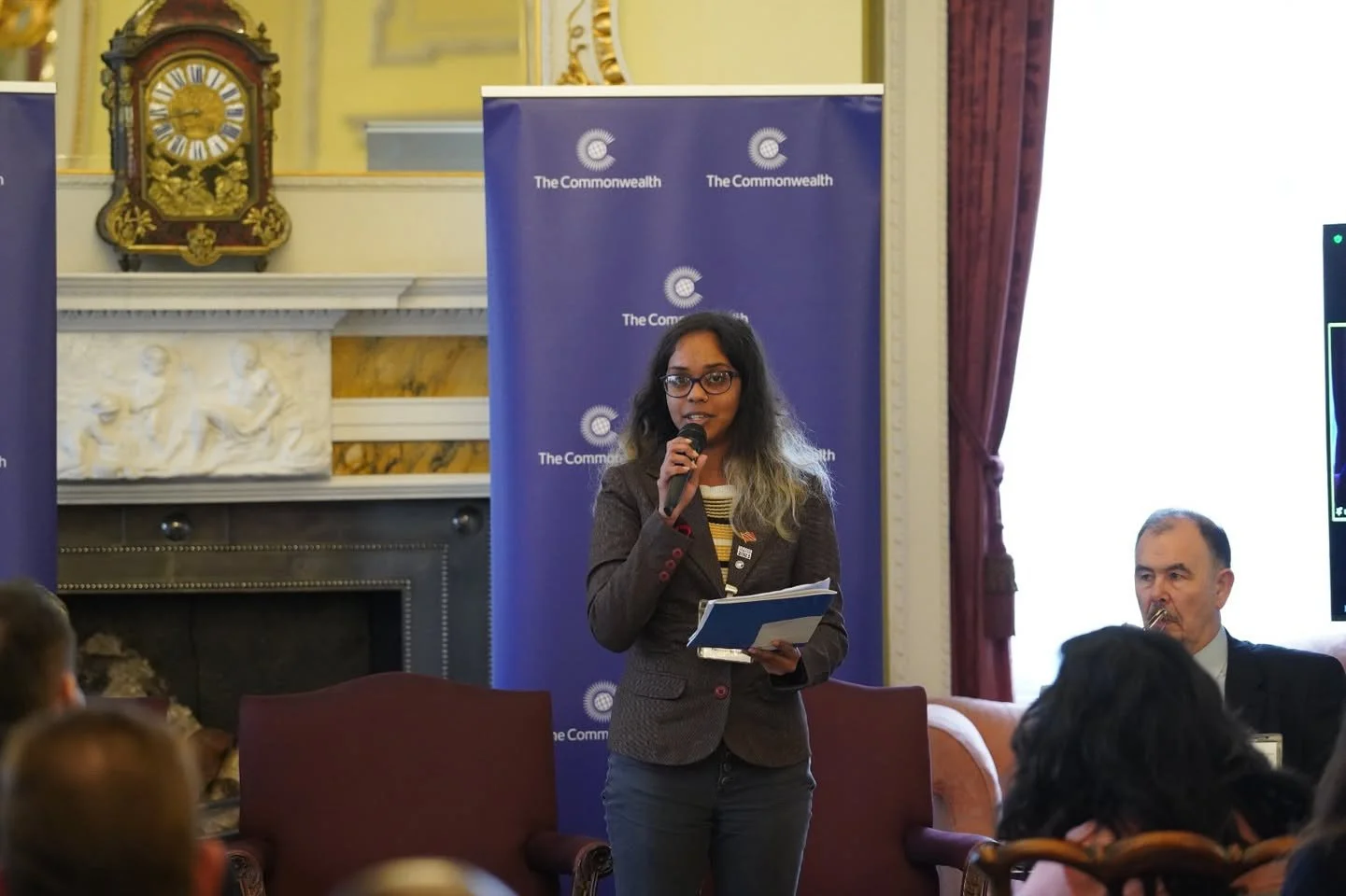 A woman with glasses and long dark hair with blonde ends is speaking into a microphone and holding a notebook. She is standing in front of a blue backdrop with 'The Commonwealth' logos. There are people seated around her, and a clock on the wall behind, along with ornate decor and curtains.