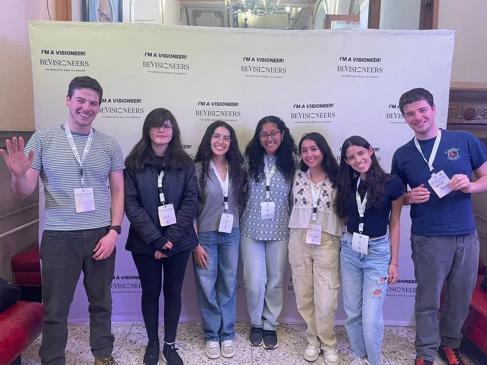 Group of seven young people standing in front of a step and repeat banner at an event, smiling and posing for a photo. The banner features the logos and slogans for BeVisioneers and the Mercedes-Benz Fellowship.