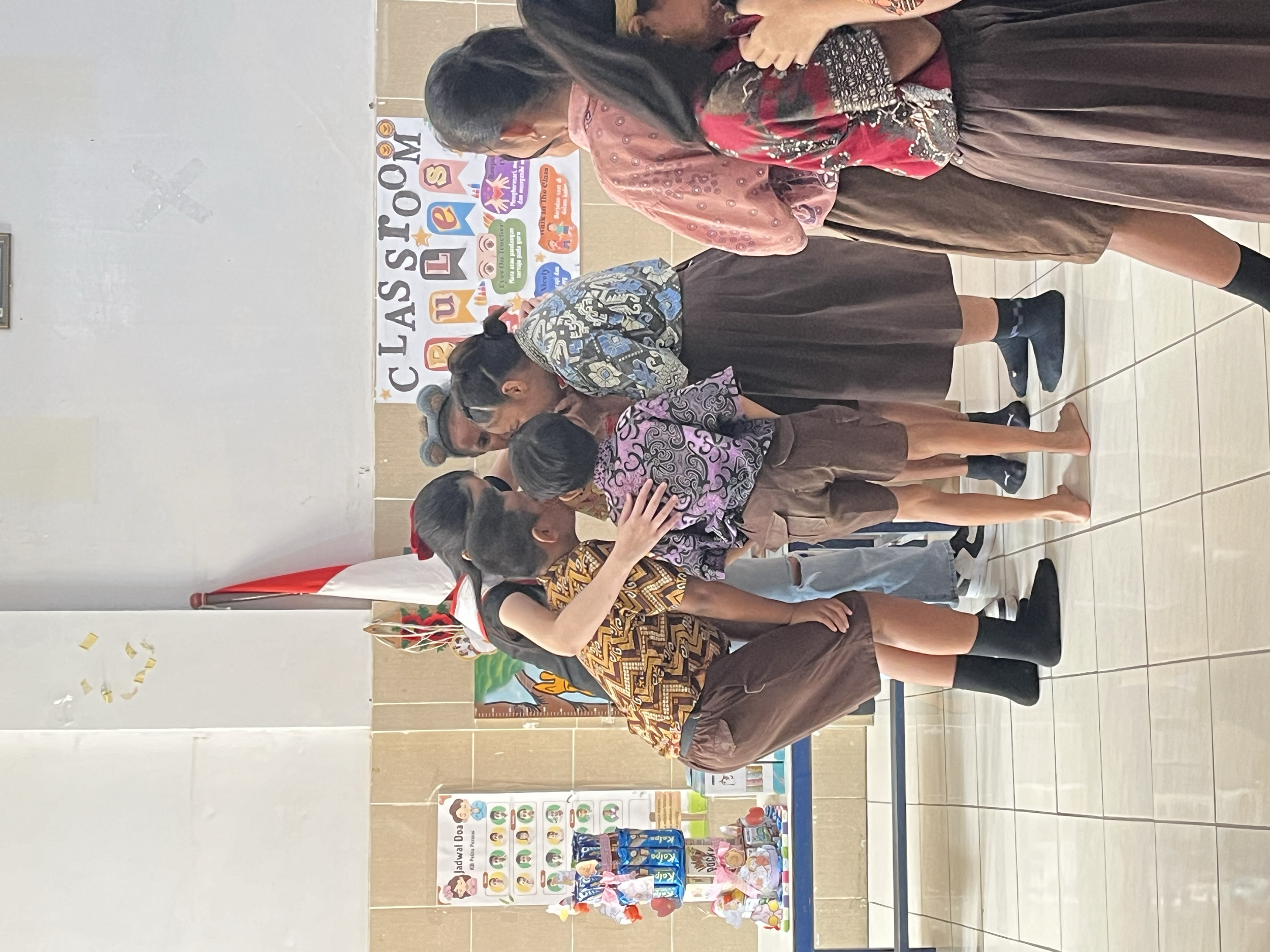 Children and adults gathered in a classroom for a Christmas celebration, with a teacher or adult dressing as Santa Claus, and a Christmas flag in the background.
