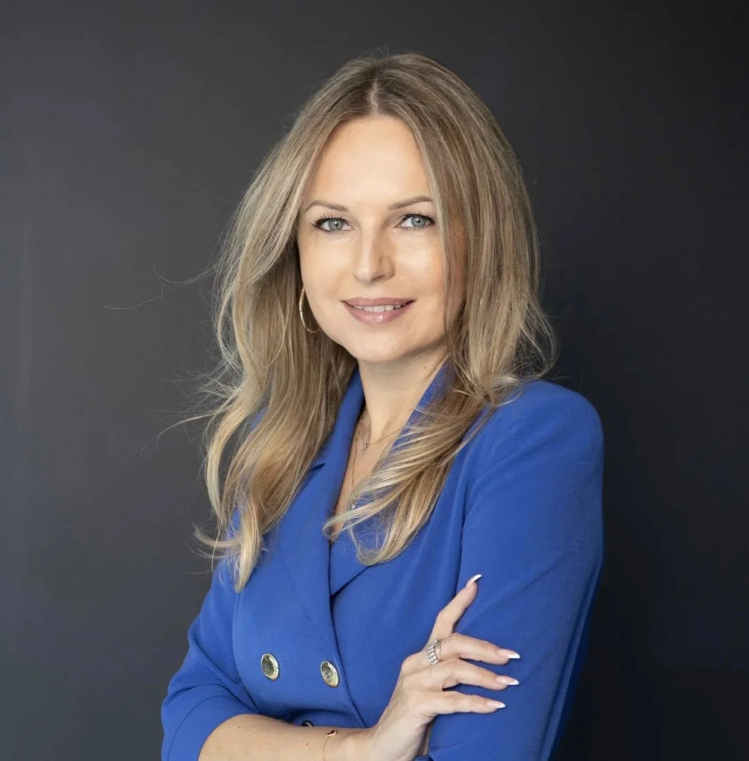 A woman with blond hair, wearing a blue blazer, with arms crossed and smiling at the camera against a dark background.