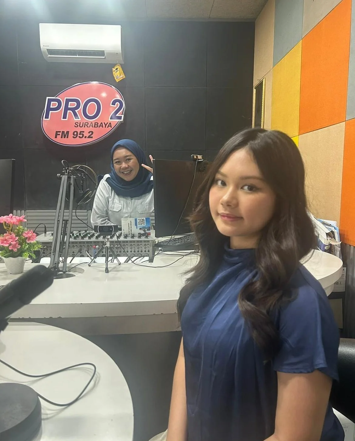 A young woman with long dark hair sitting in a radio studio, with a woman in a hijab and white shirt smiling and talking behind a radio console. The studio has a sign that reads 'PRO 2 SURABAYA FM 95.2' on the wall.
