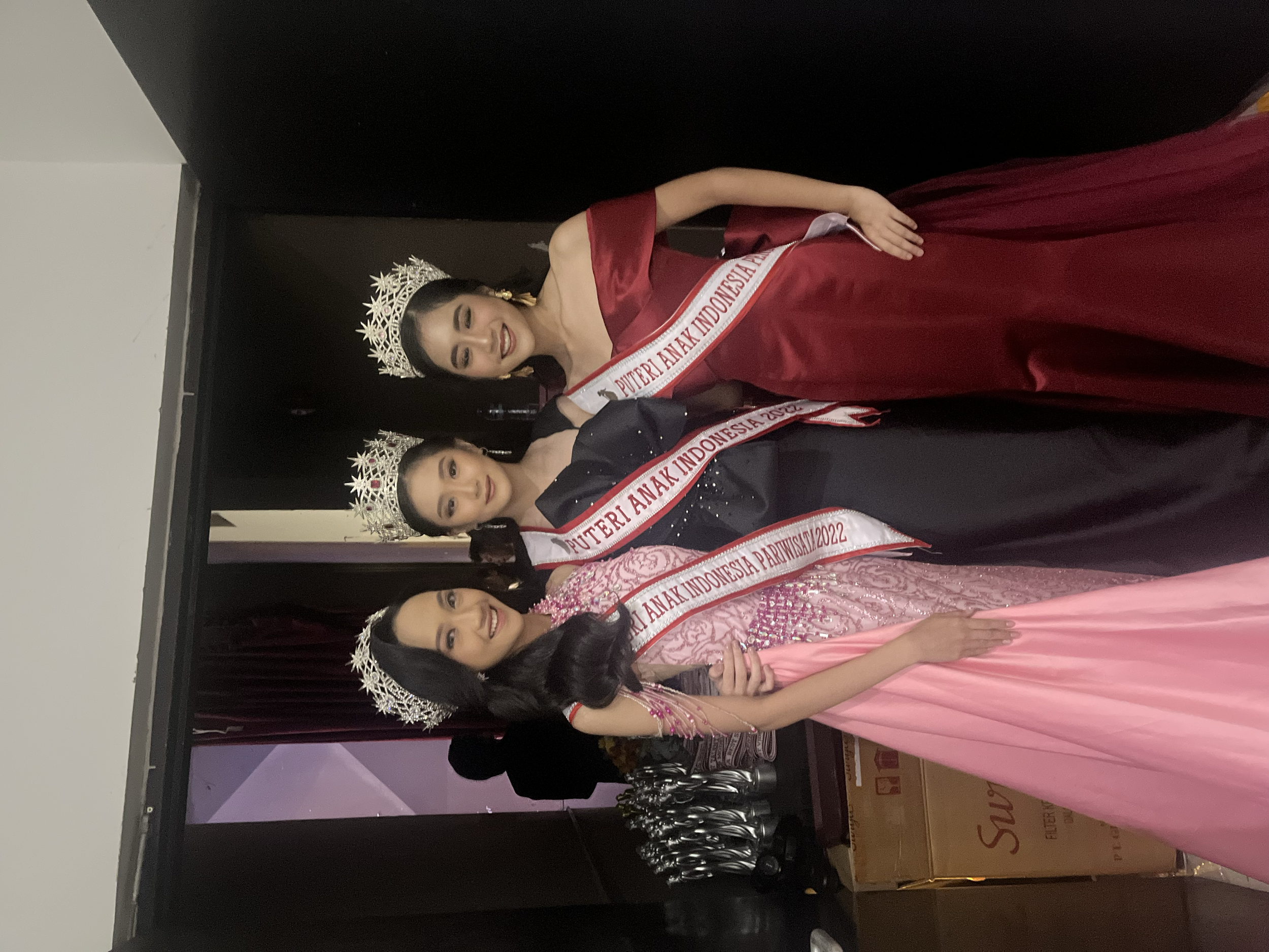 Three women wearing evening gowns and crowns, each with a sash, standing on a stage or backstage area. They are smiling and posing for a photo together.